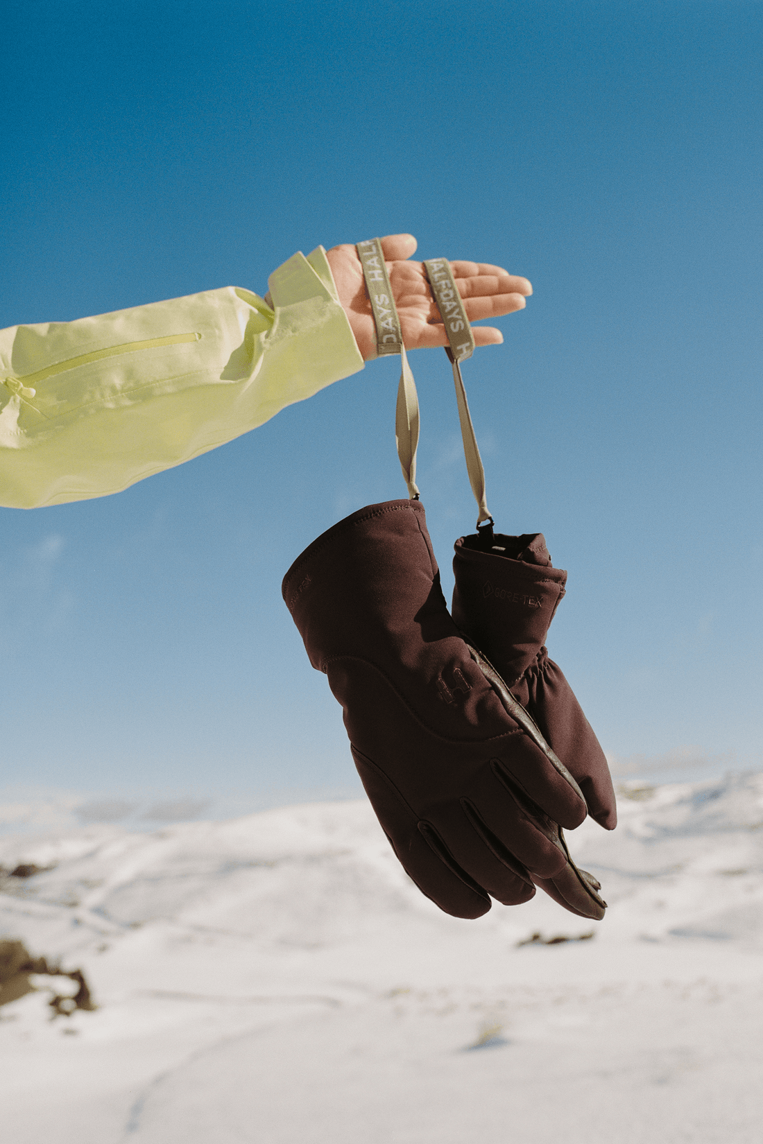 A pair of dark brown insulated gloves hanging from a person's hand, who is wearing a pale yellow jacket, set against a snowy mountain landscape and clear blue sky.