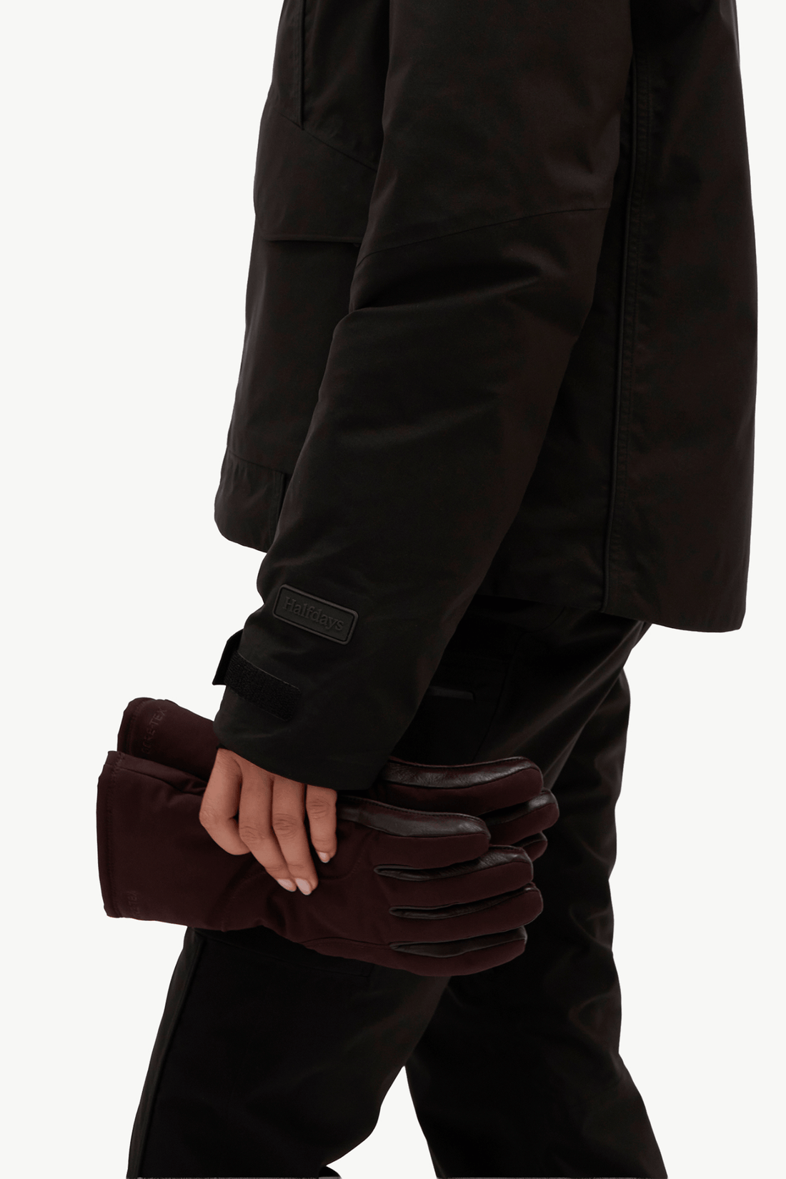 A close-up of a person wearing a black jacket and black pants, holding a pair of dark burgundy gloves against a plain light background.