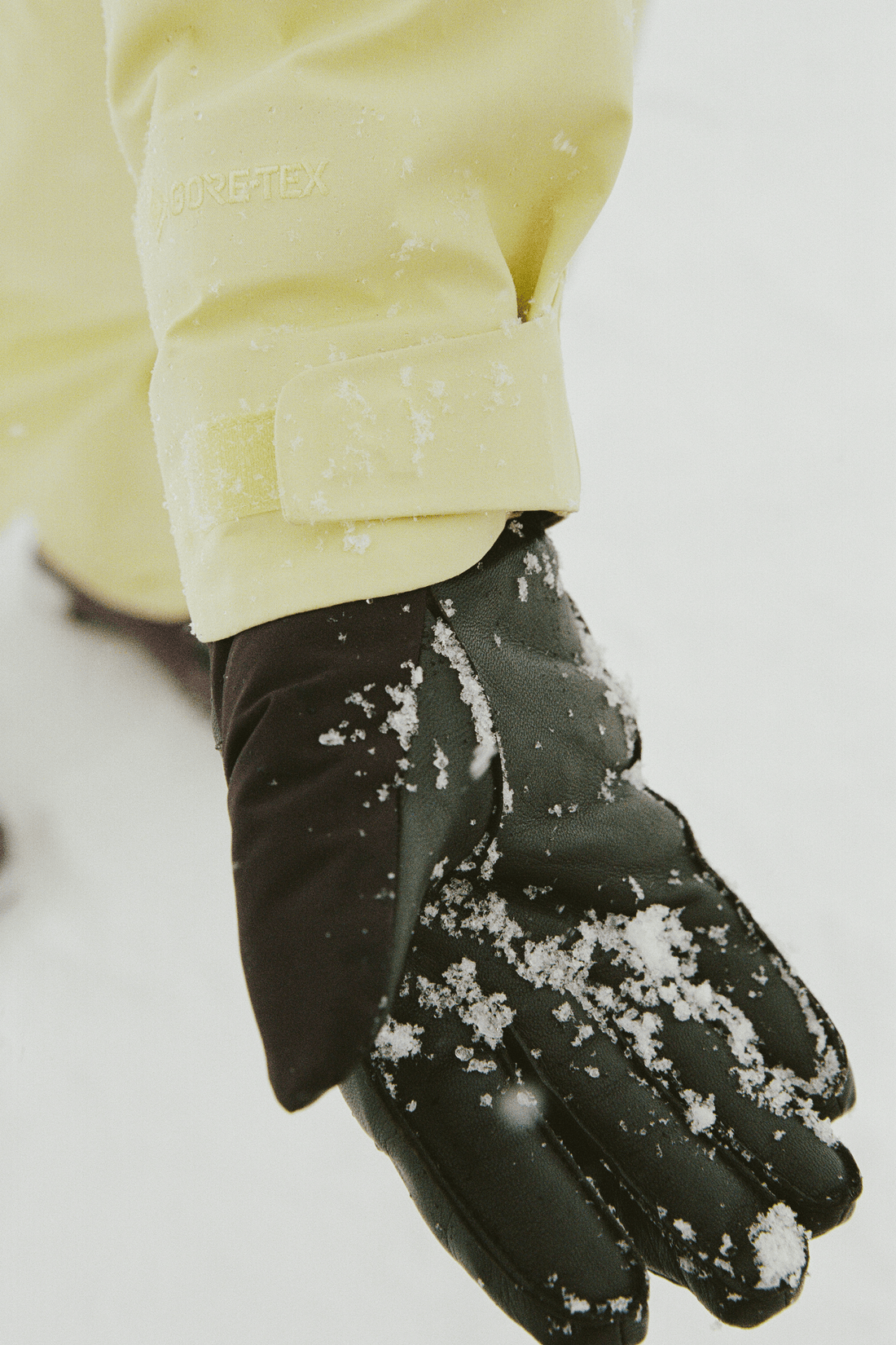 A close-up of a black winter glove covered in snow, worn with a pale yellow jacket sleeve, against a snowy white background.
