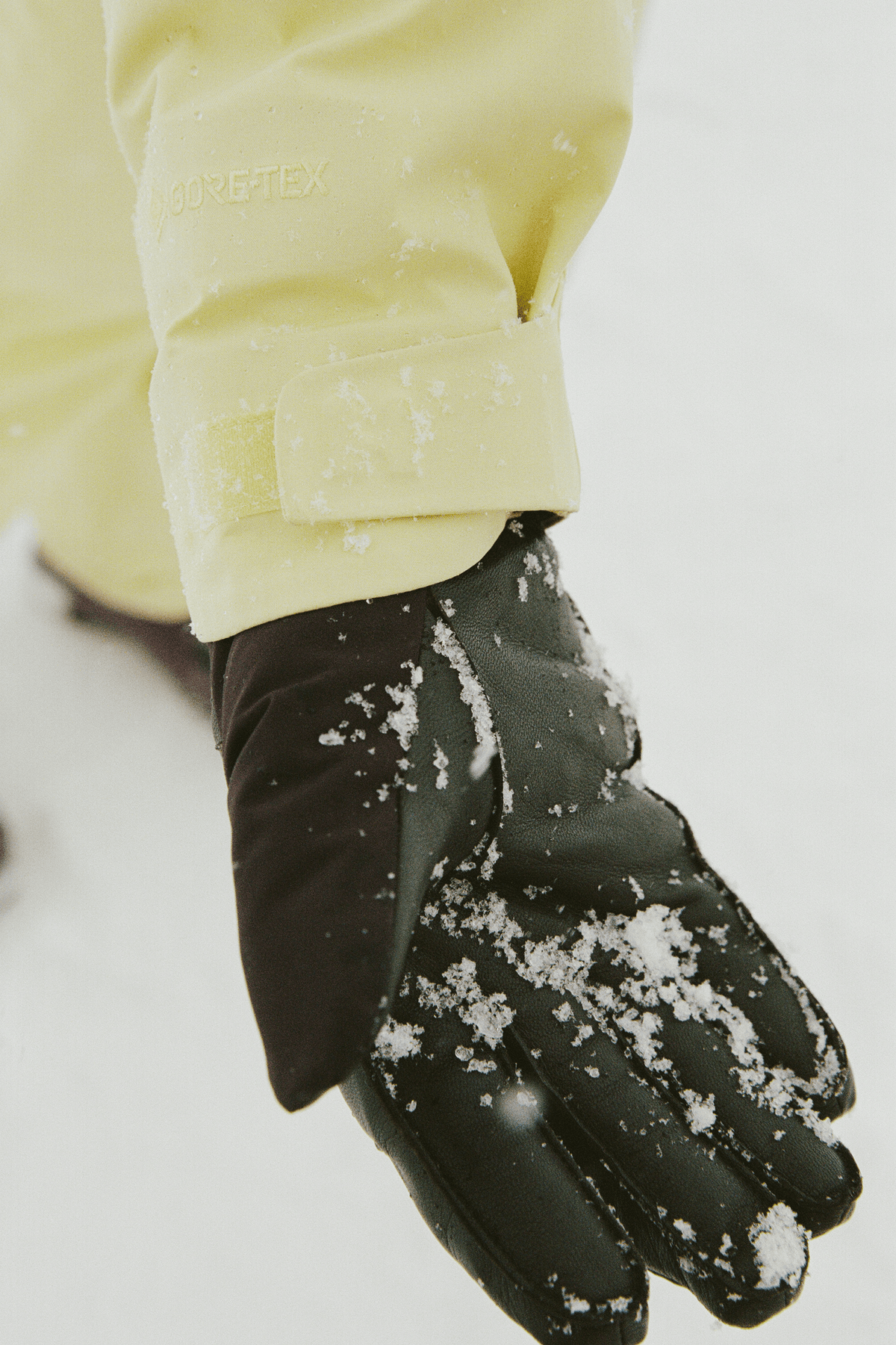 A close-up of a black winter glove covered in snow, worn with a pale yellow jacket sleeve, against a snowy white background.