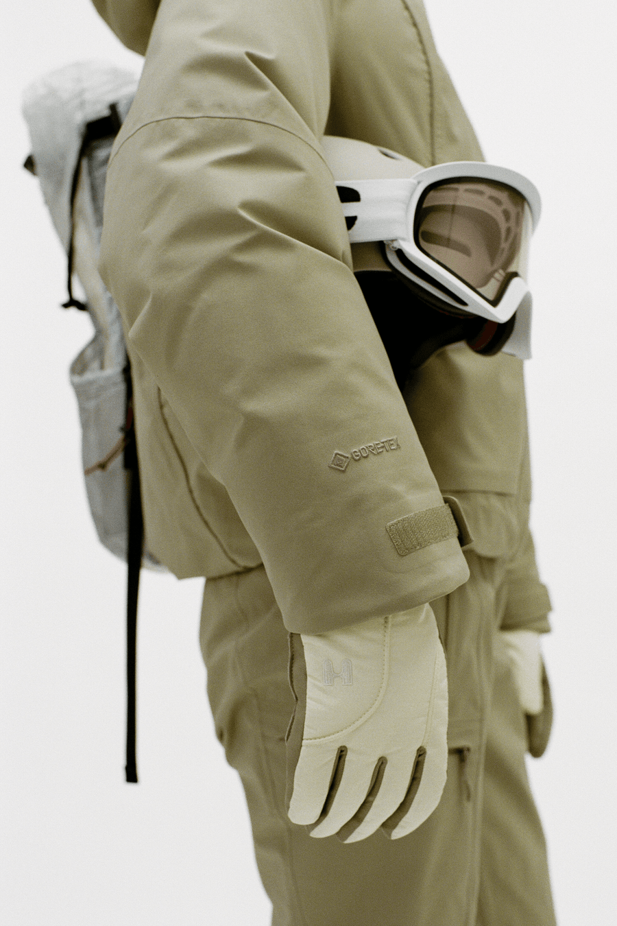 A close-up of a person wearing a beige technical ski jacket and matching pants, with off-white gloves and white ski goggles resting on the jacket. A small Darn Tough logo is visible on the sleeve.
