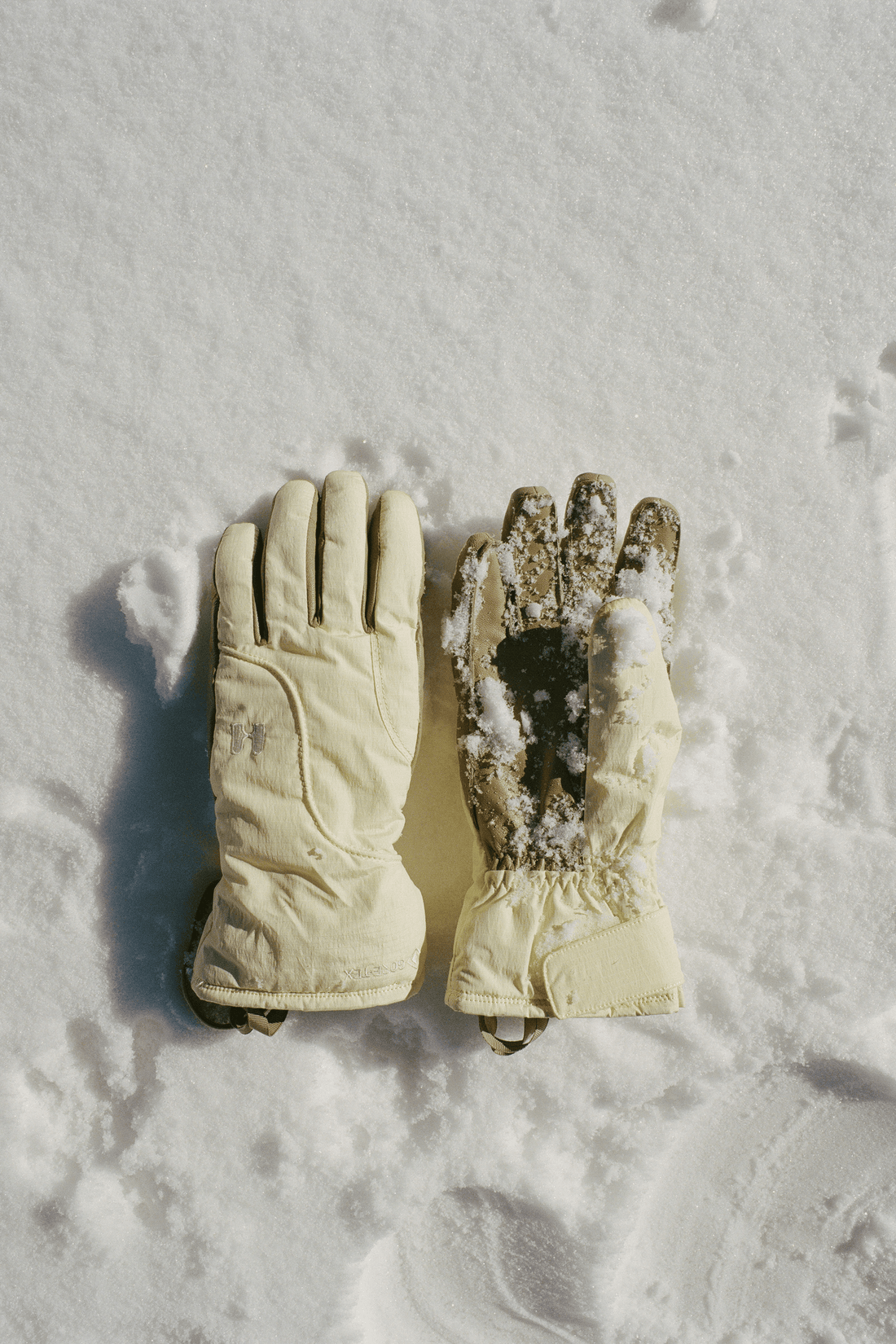 A pair of off-white insulated winter gloves placed on snow, with one glove showing the back and the other showing the palm side dusted with snow.