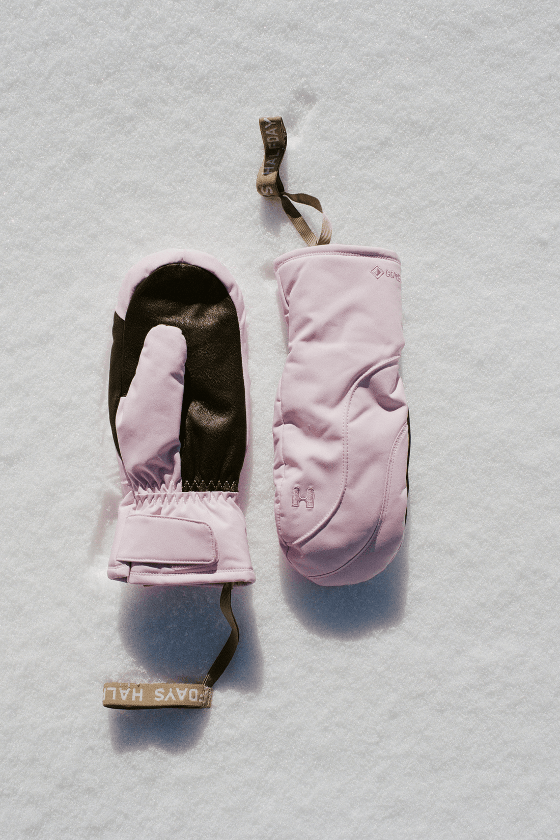 A pair of light pink winter mittens laid flat on a white snowy surface. One mitten is shown open, revealing a dark brown interior, and the other is closed, showing a pale pink exterior with a small tonal logo.
