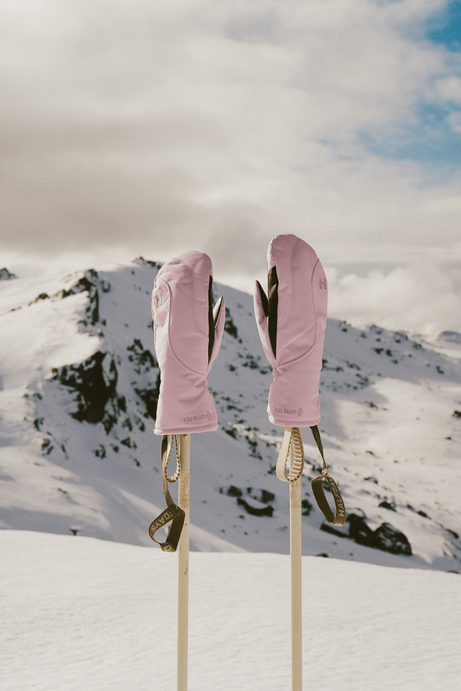 A pair of light pink winter mittens hanging from ski poles in a snowy mountain landscape under a cloudy sky.