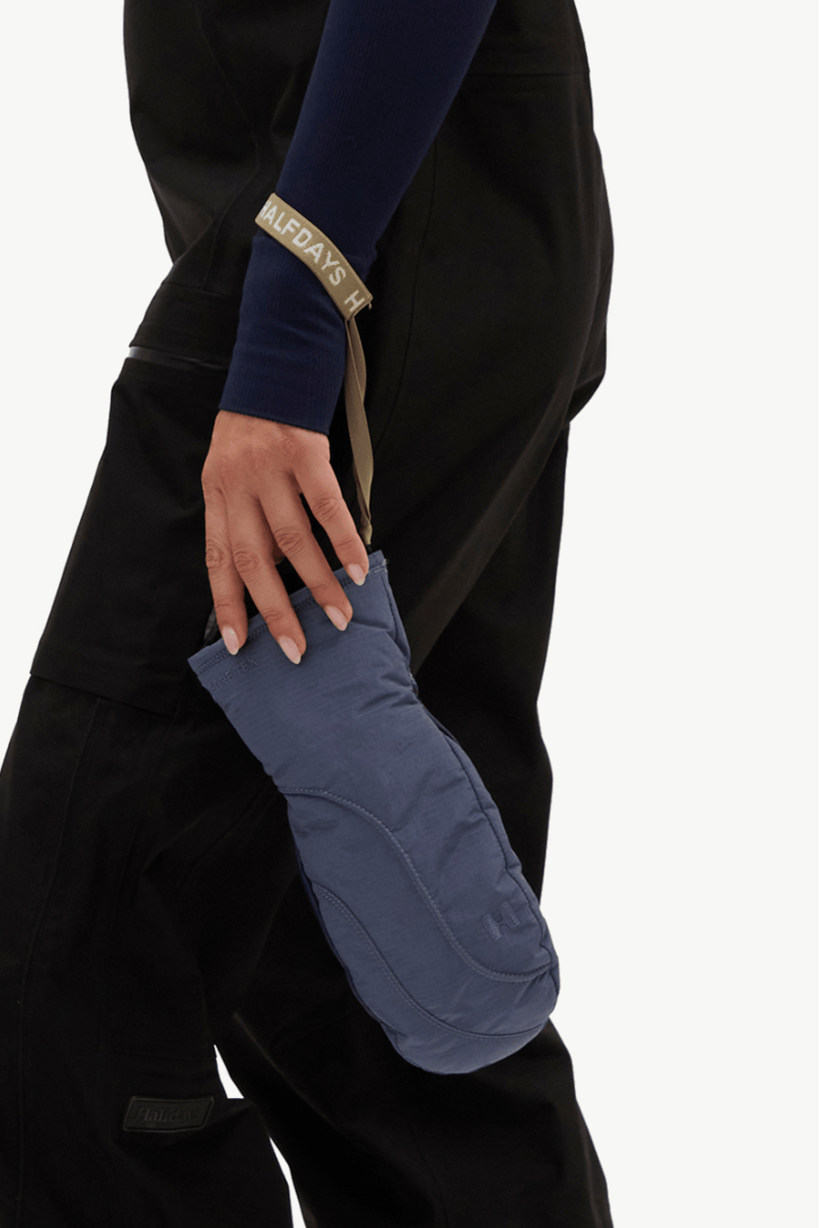 A close-up of a person wearing black outdoor pants and a navy long-sleeve top, holding a slate blue insulated glove or puffer sleeve. A tan strap with black text is attached near the cuff.