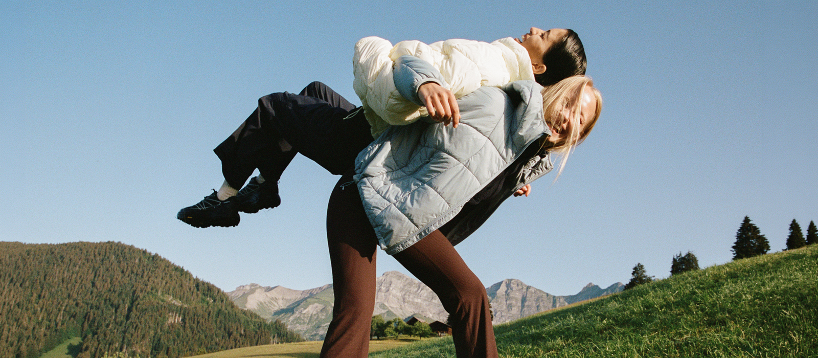 Two people wearing Halfdays outerwear playfully posing in a grassy mountain landscape under a clear blue sky.