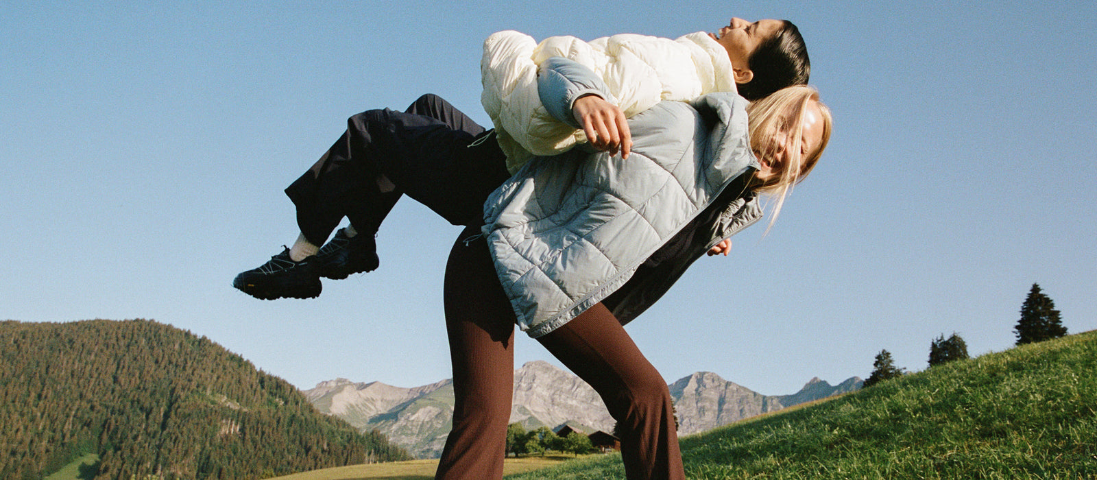 Two people wearing Halfdays outerwear playfully posing in a grassy mountain landscape under a clear blue sky.