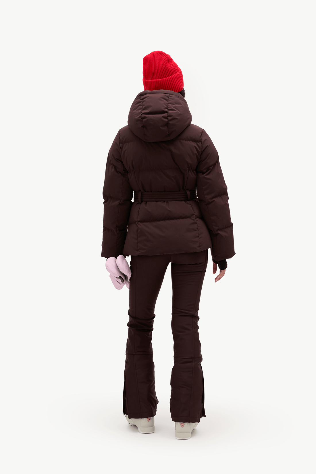 A model shown from the back wearing a dark brown puffer jacket with a matching belt, dark brown flared pants, a bright red knit beanie, and light pink gloves, set against a plain white studio background.