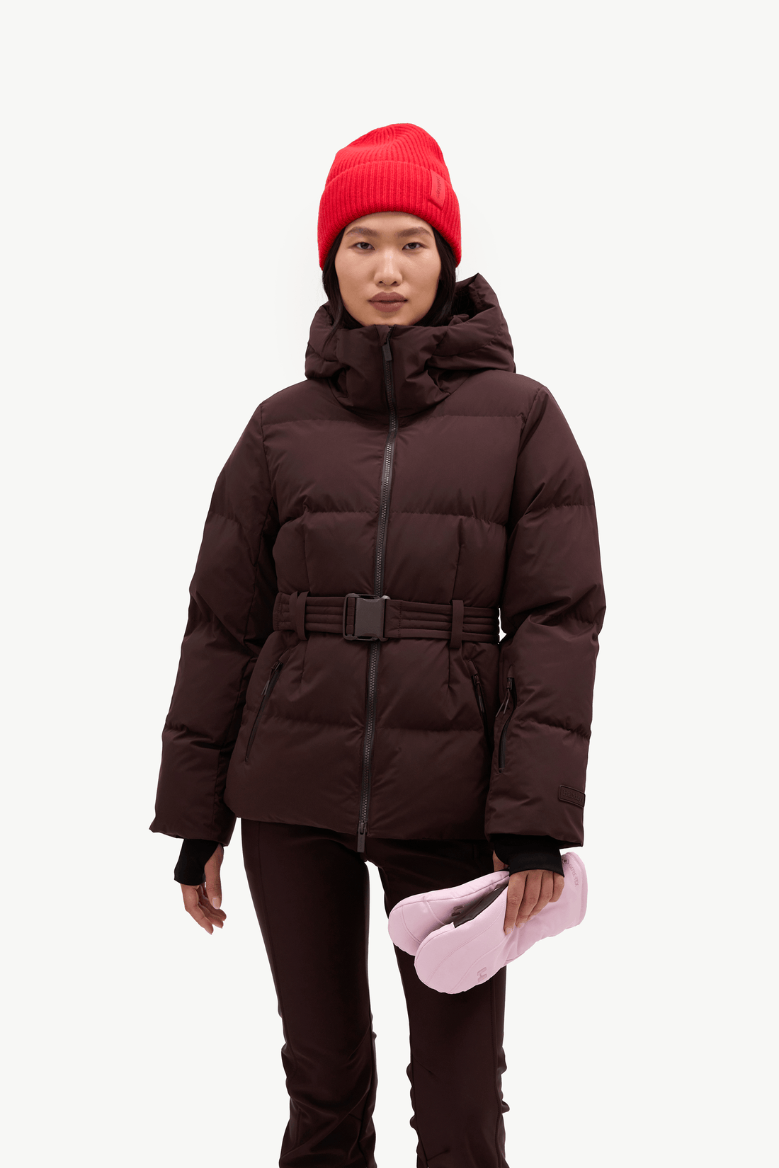 A model wearing a dark brown puffer jacket with a matching belt, dark brown pants, a bright red knit beanie, and holding a pair of light pink winter boots against a plain white background.