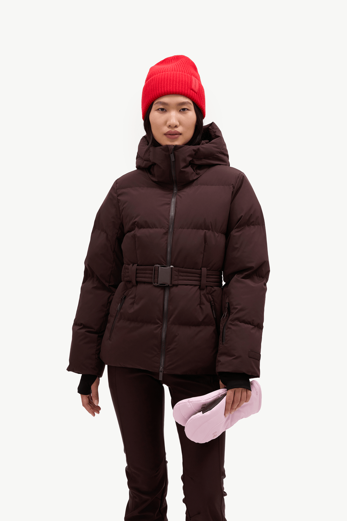 A model wearing a dark brown puffer jacket with a matching belt, dark brown pants, a bright red knit beanie, and holding a pair of light pink winter boots against a plain white background.