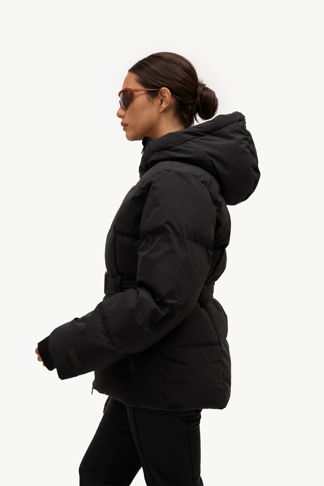 A woman in profile wearing a black puffer jacket with a hood and a matching black belt, photographed against a clean white studio background.