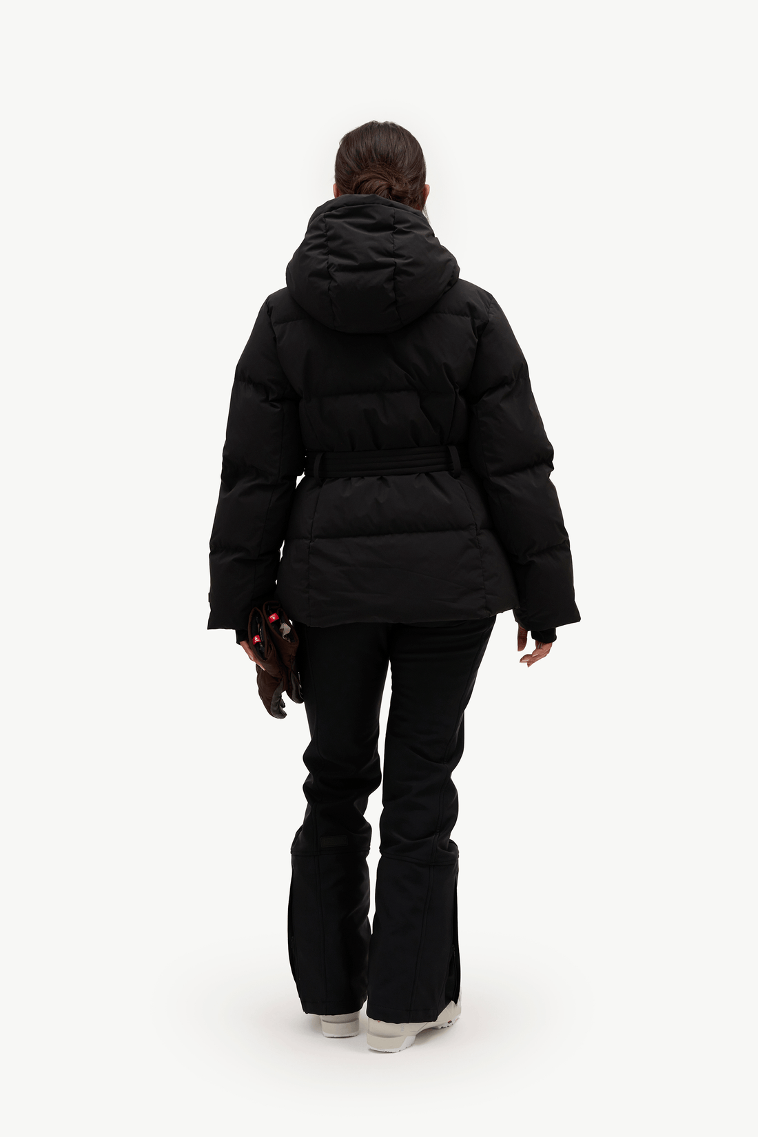 A model stands against a plain white studio background wearing a black hooded puffer jacket with a front zipper, a matching black belt, black pants, and light gray sneakers. She is holding a pair of dark brown gloves.