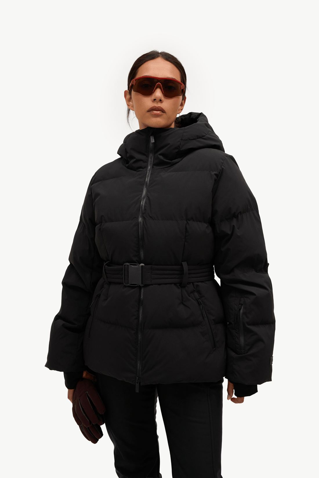 A model stands against a plain white studio background wearing a black hooded puffer jacket with a front zipper, a matching black belt, black pants, and light gray sneakers. She is holding a pair of dark brown gloves.