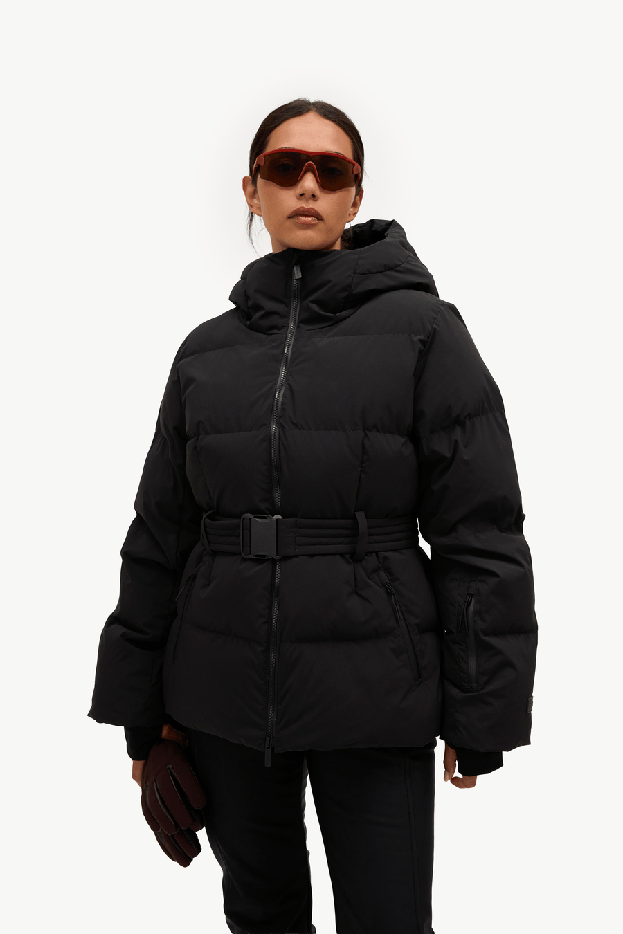 A model stands against a plain white studio background wearing a black hooded puffer jacket with a front zipper, a matching black belt, black pants, and light gray sneakers. She is holding a pair of dark brown gloves.