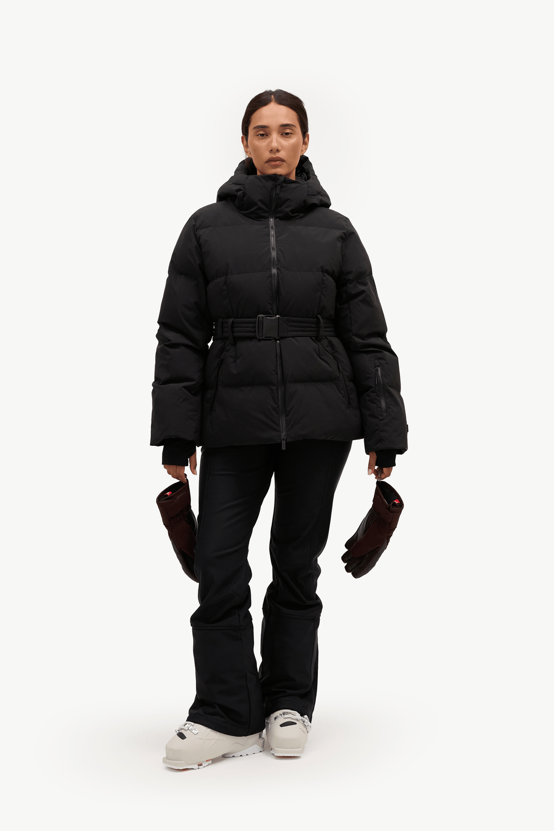 A model stands against a plain white studio background wearing a black hooded puffer jacket with a front zipper, a matching black belt, black pants, and light gray sneakers. She is holding a pair of dark brown gloves.