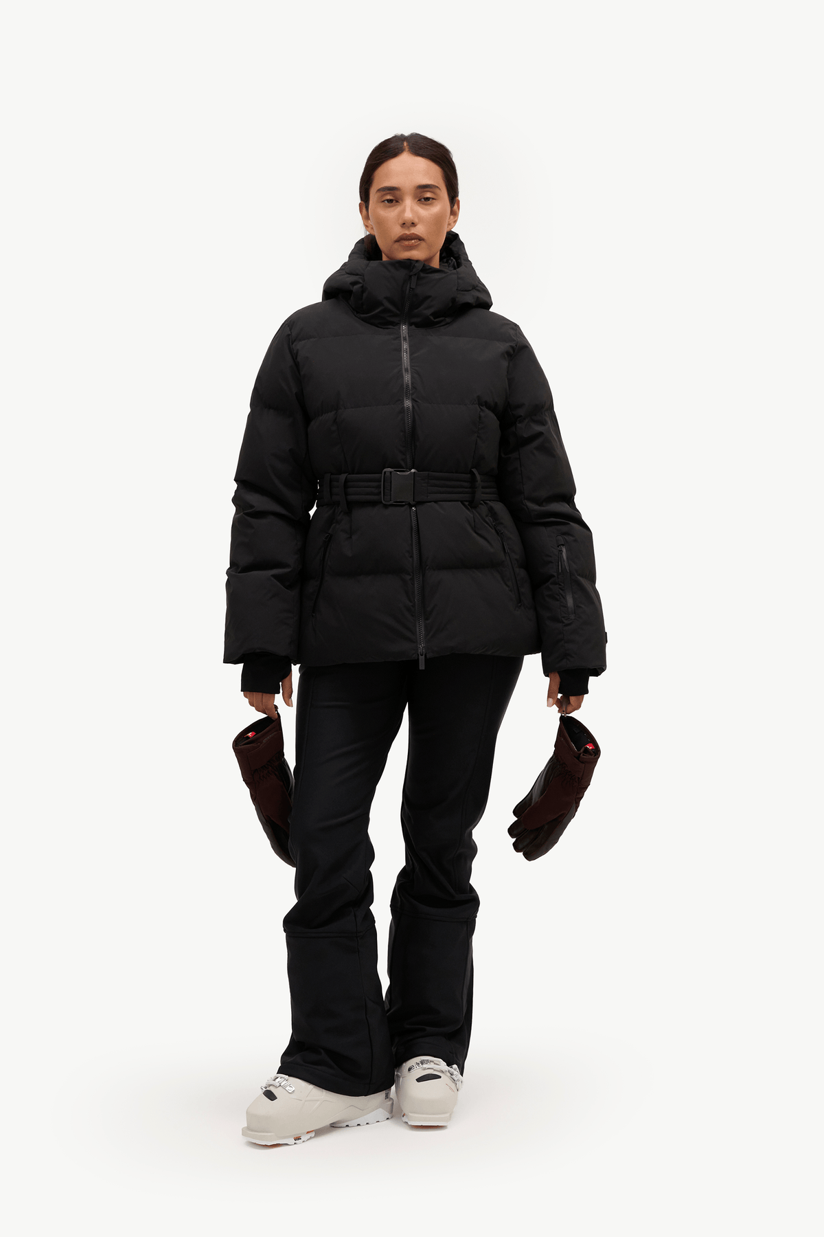 A model stands against a plain white studio background wearing a black hooded puffer jacket with a front zipper, a matching black belt, black pants, and light gray sneakers. She is holding a pair of dark brown gloves.
