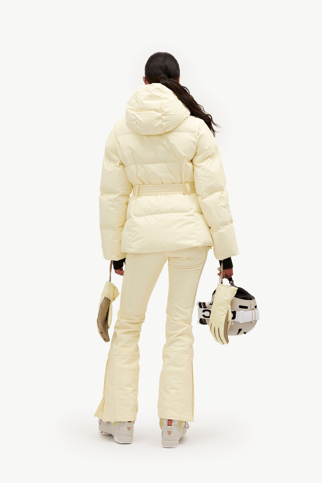 A model shown from the back wearing a cream-colored puffer jacket and matching flared pants, holding a ski helmet and gloves against a plain white background.