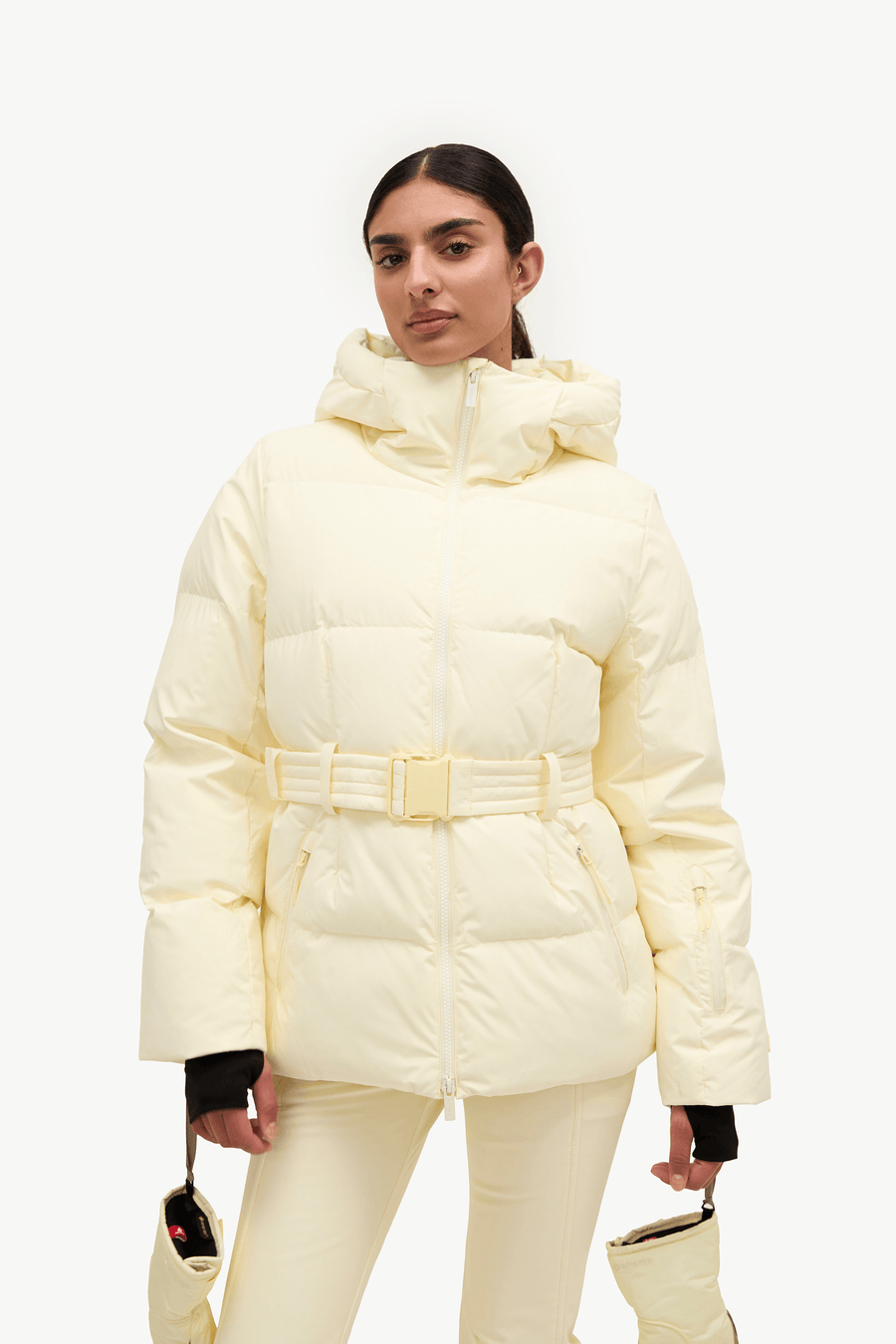 A model wearing a yellow-colored belted puffer jacket with a matching hood, paired with matching cream pants and holding matching cream handbags against a plain white background.