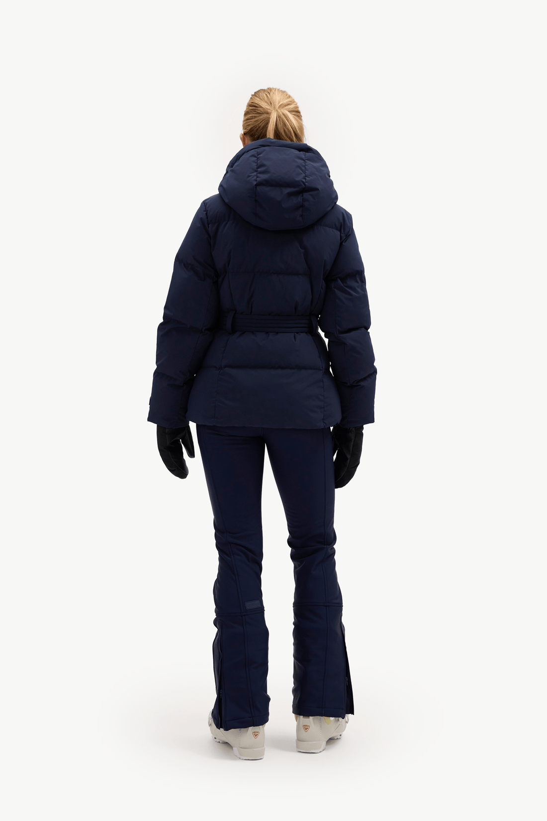 A woman modeling a dark navy blue belted puffer jacket, matching navy pants, black gloves, and white sneakers against a plain white studio background.