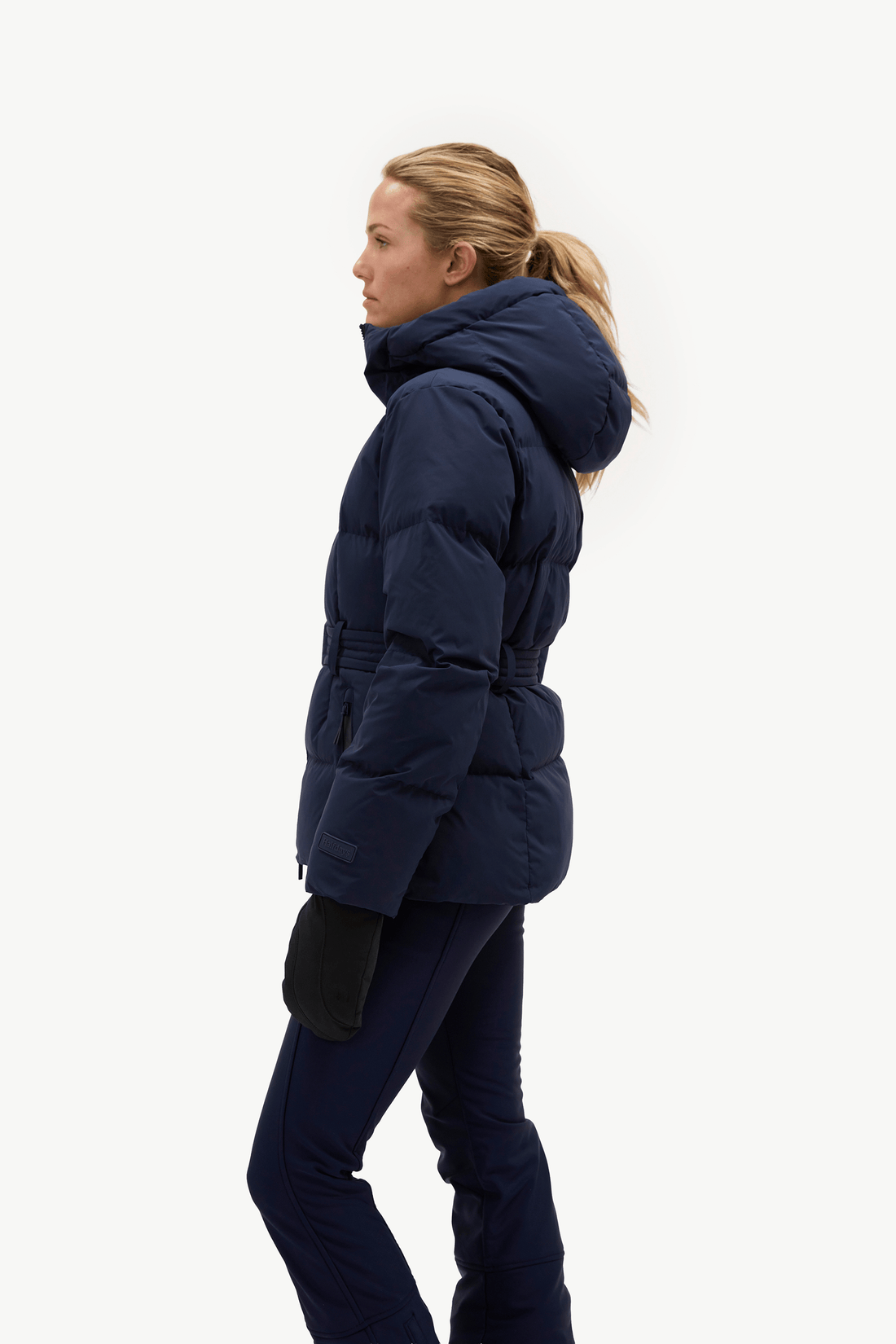 A woman in side profile wearing a navy blue puffer jacket with a high collar and a matching navy blue pair of pants, standing against a solid white background.