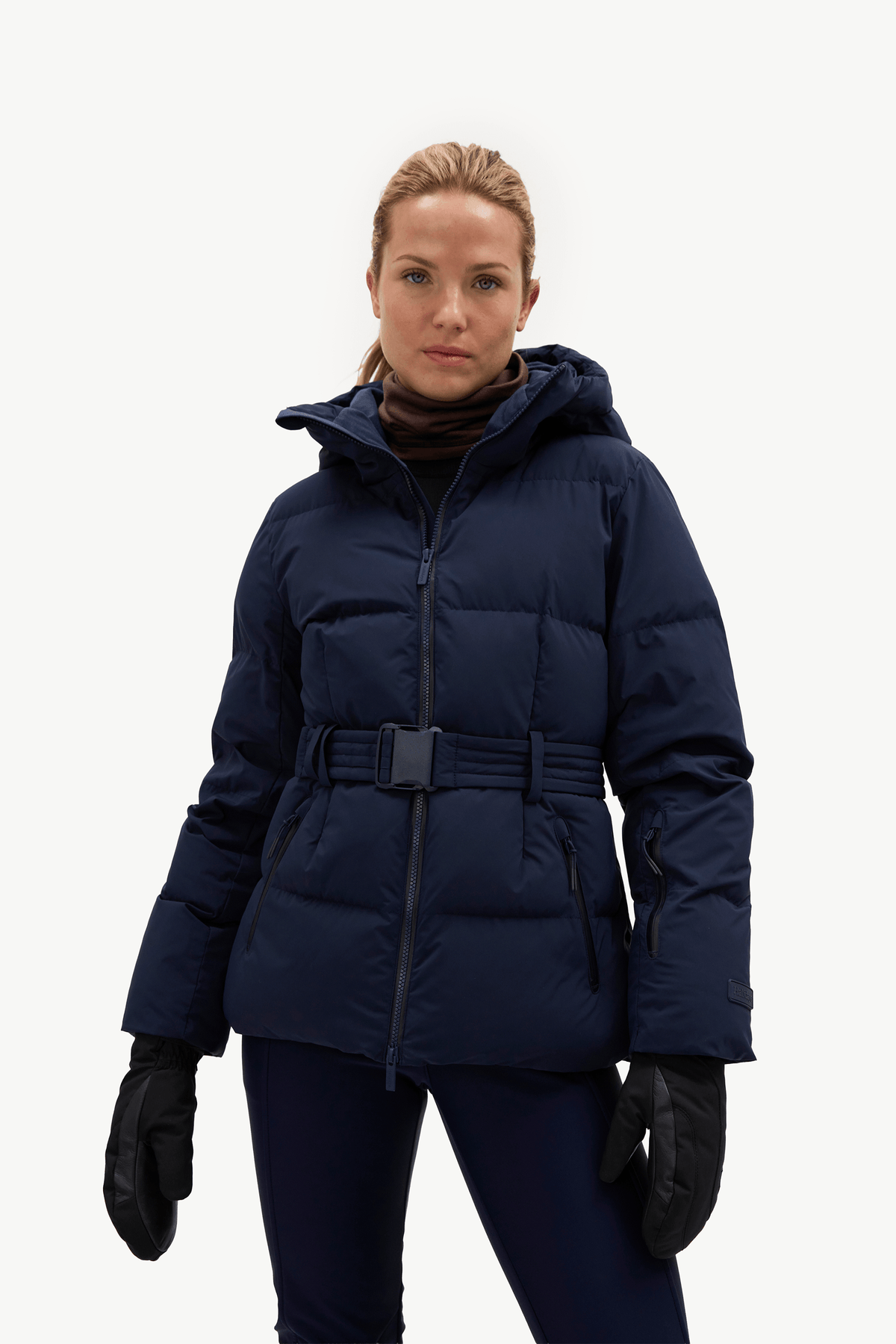 A woman modeling a dark navy blue belted puffer jacket, matching navy pants, black gloves, and white sneakers against a plain white studio background.