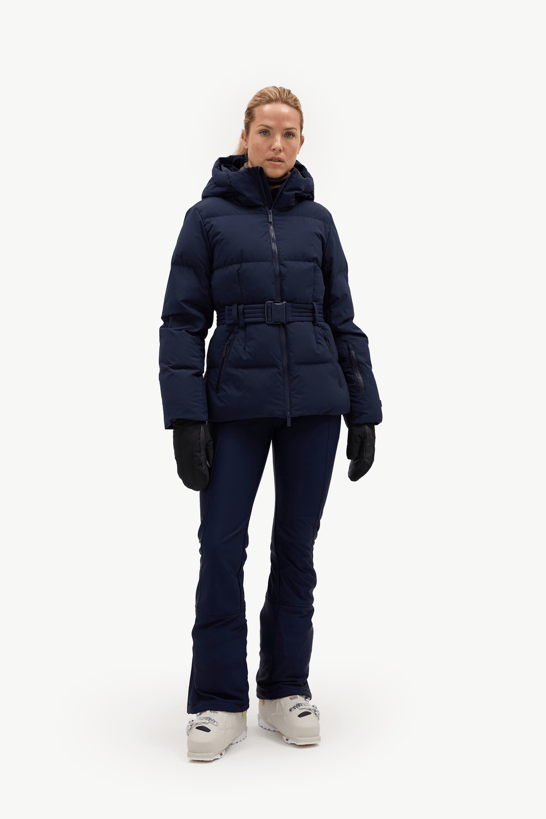A woman modeling a dark navy blue belted puffer jacket, matching navy pants, black gloves, and white sneakers against a plain white studio background.