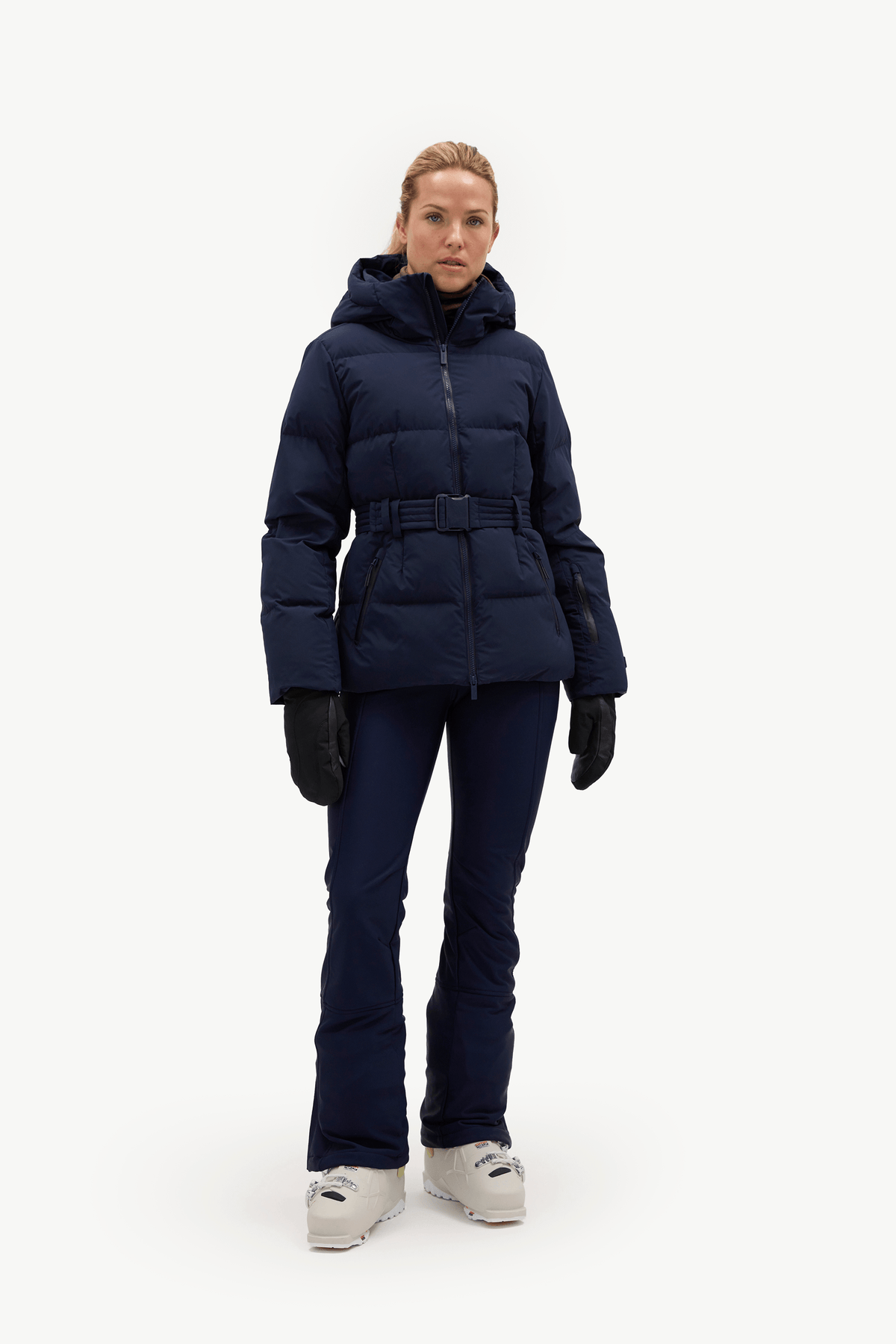 A woman modeling a dark navy blue belted puffer jacket, matching navy pants, black gloves, and white sneakers against a plain white studio background.