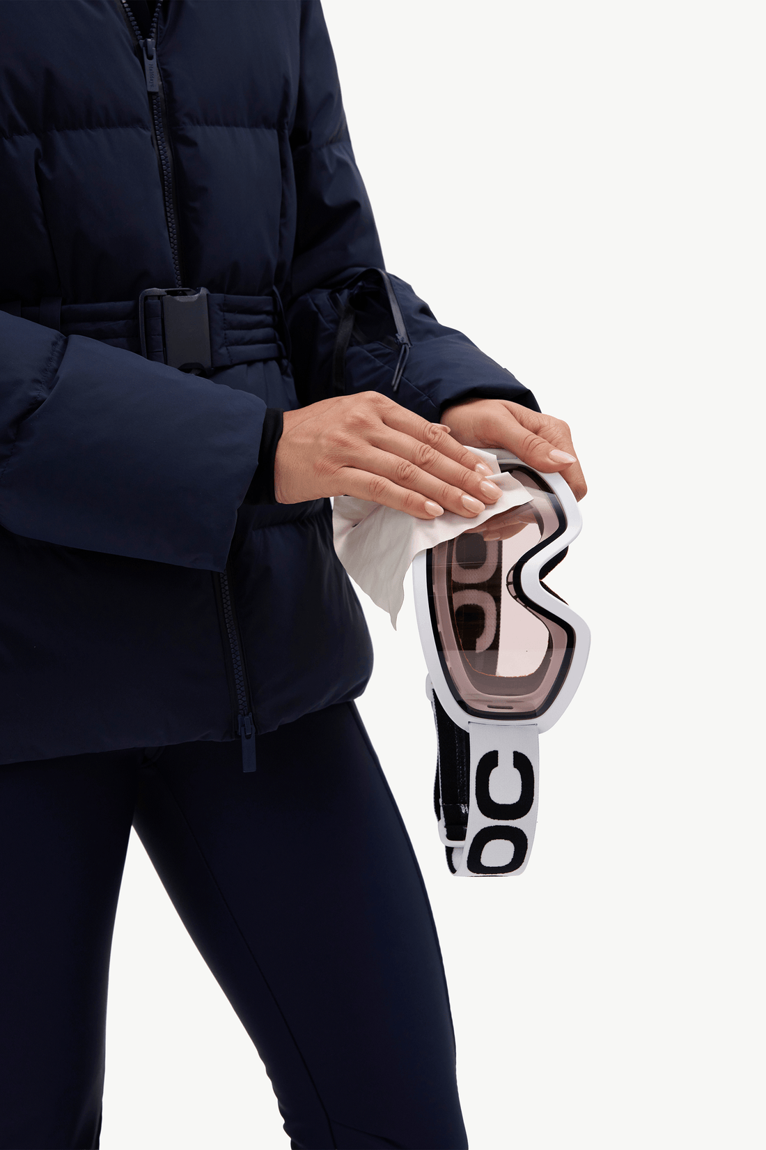 A close-up of a person in a navy puffer jacket holding a pair of ski goggles with a white cloth, against a clean white background.
