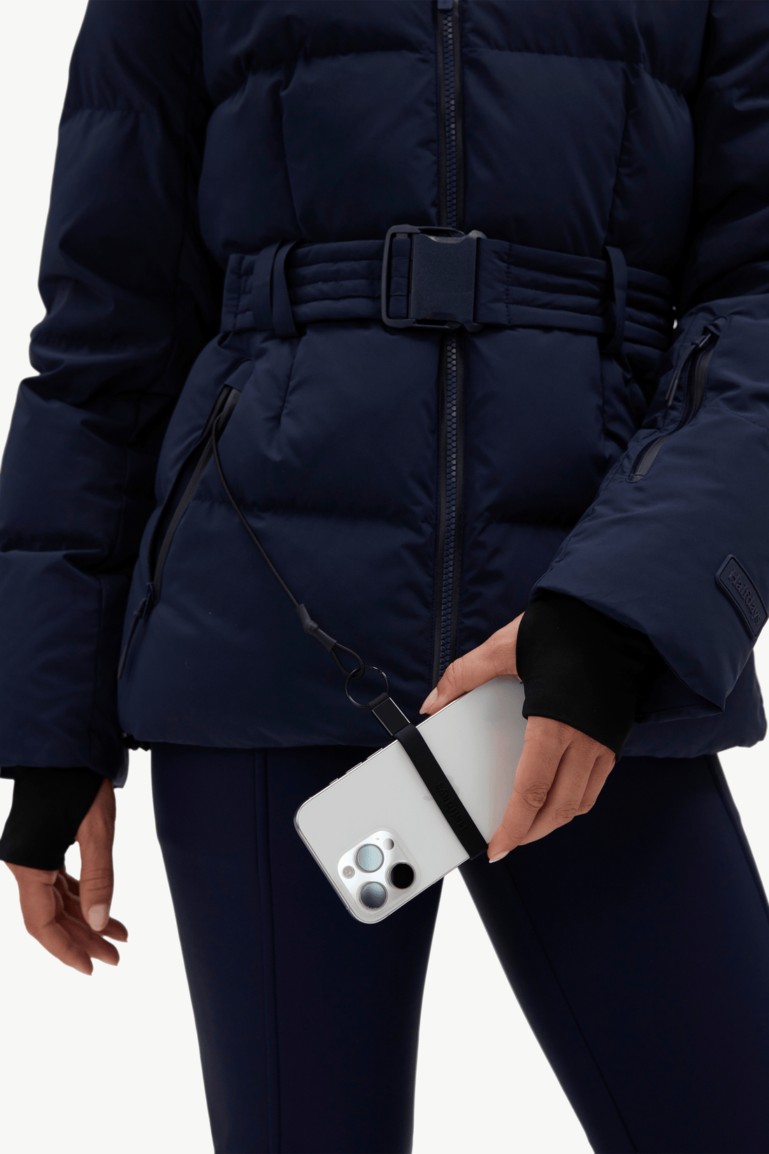 A close-up of a person wearing a navy blue puffer jacket with a matching belt and dark leggings, holding a white smartphone with a black crossbody strap attached.