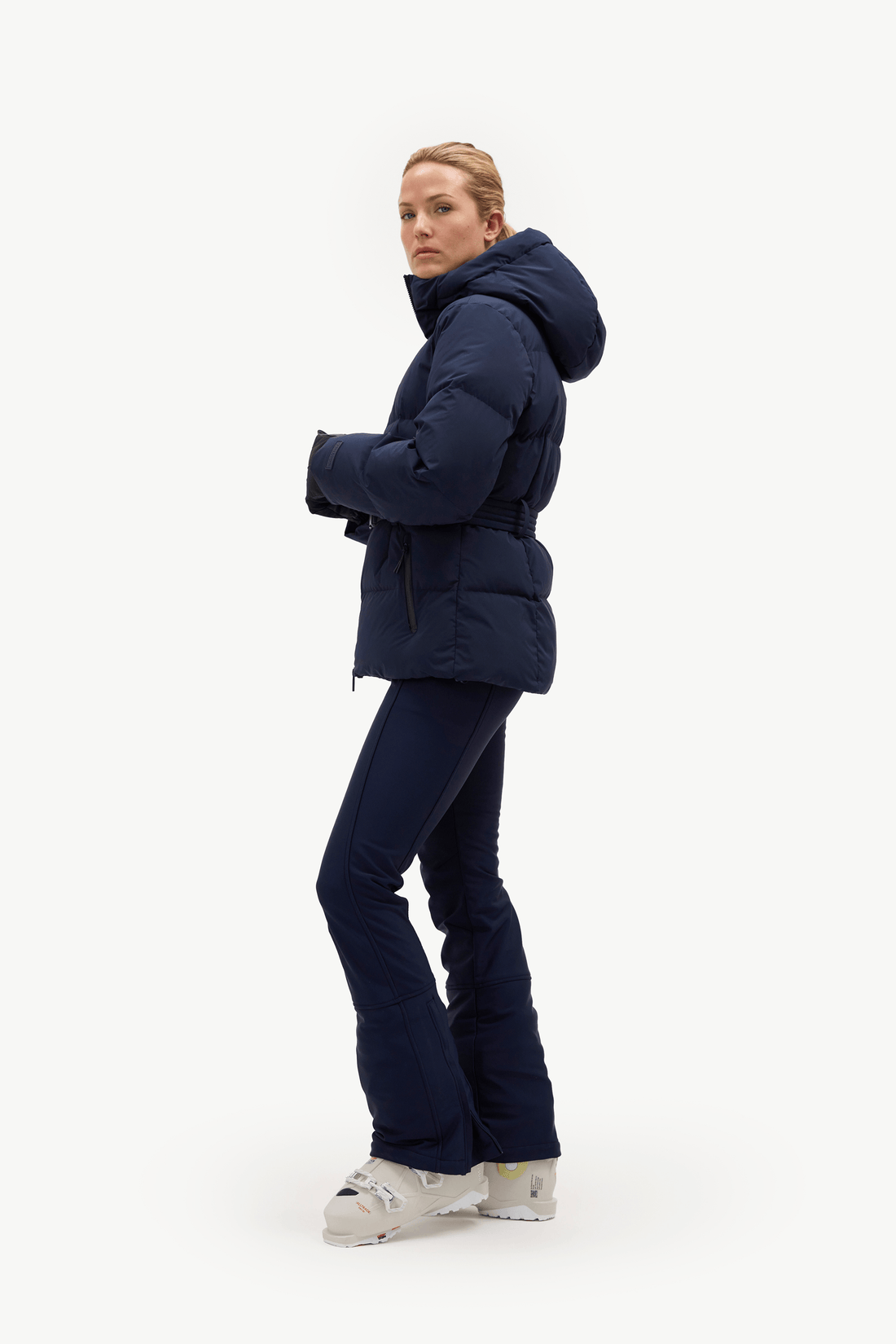 A woman modeling a navy blue hooded puffer jacket and matching navy ski pants with white ski boots, standing against a solid white studio background.