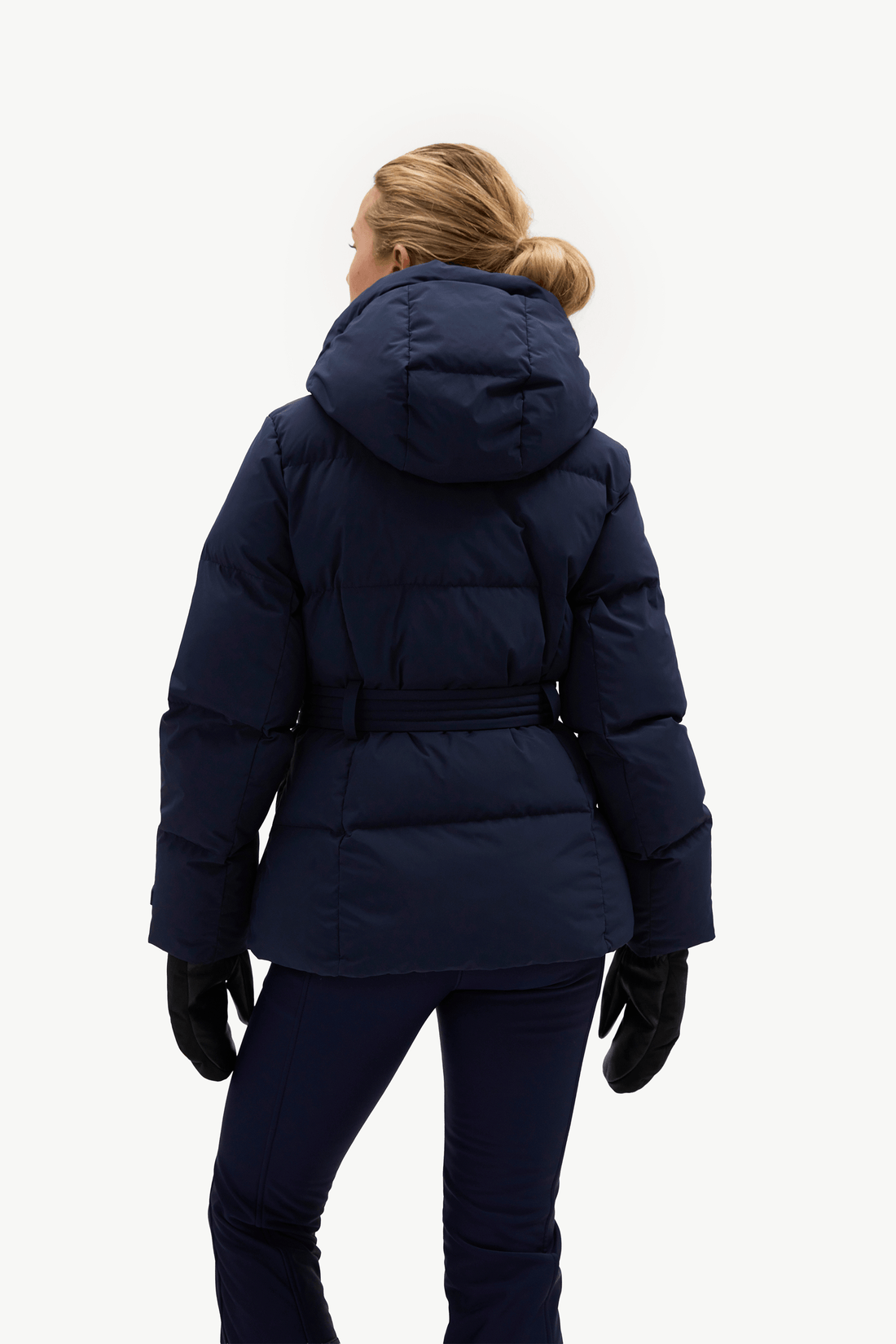 A woman modeling a dark navy blue belted puffer jacket, matching navy pants, black gloves, against a plain white studio background.
