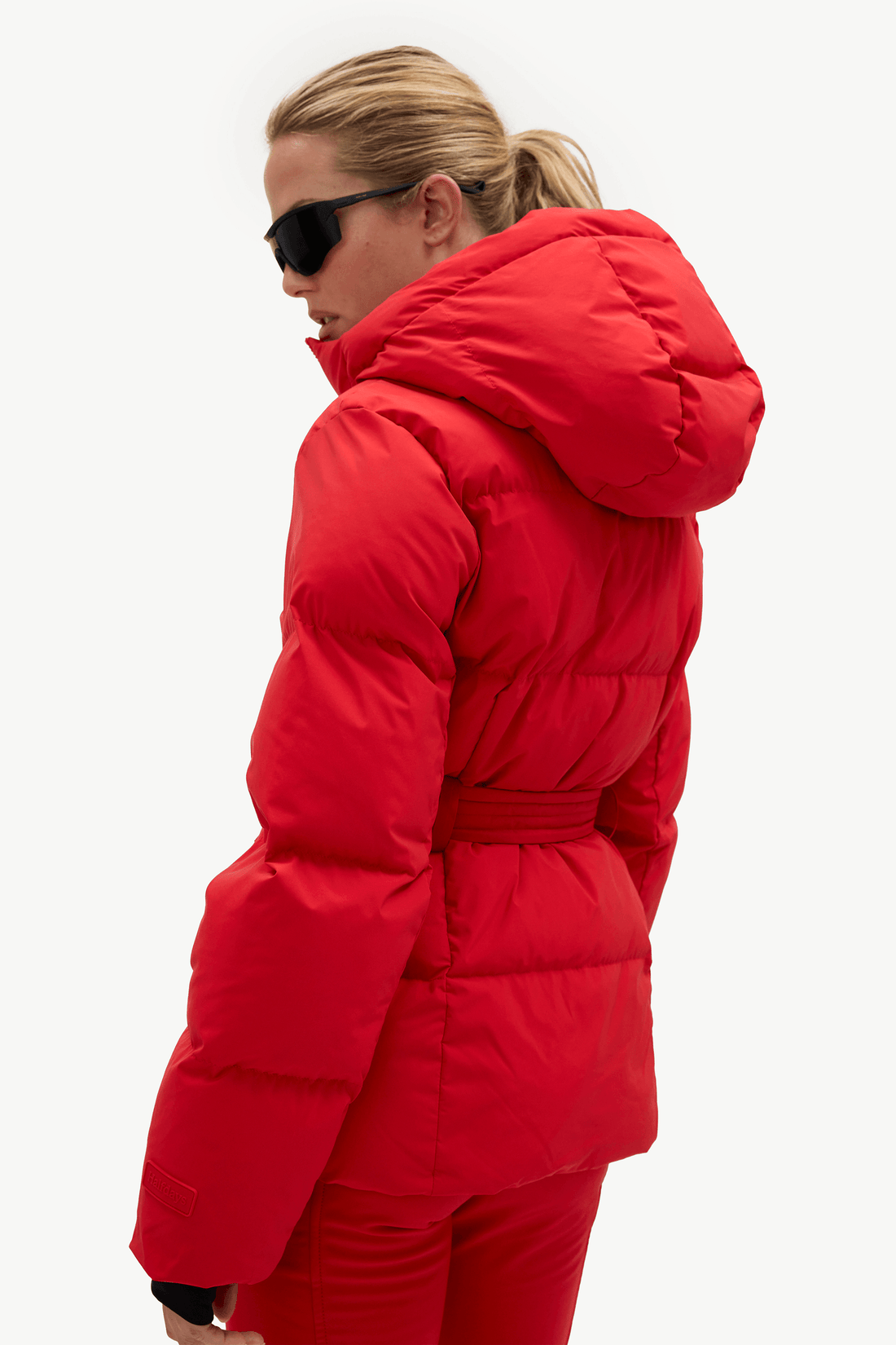 A woman wearing a bright red hooded puffer jacket with a matching belt and red pants, photographed against a plain light gray studio background.