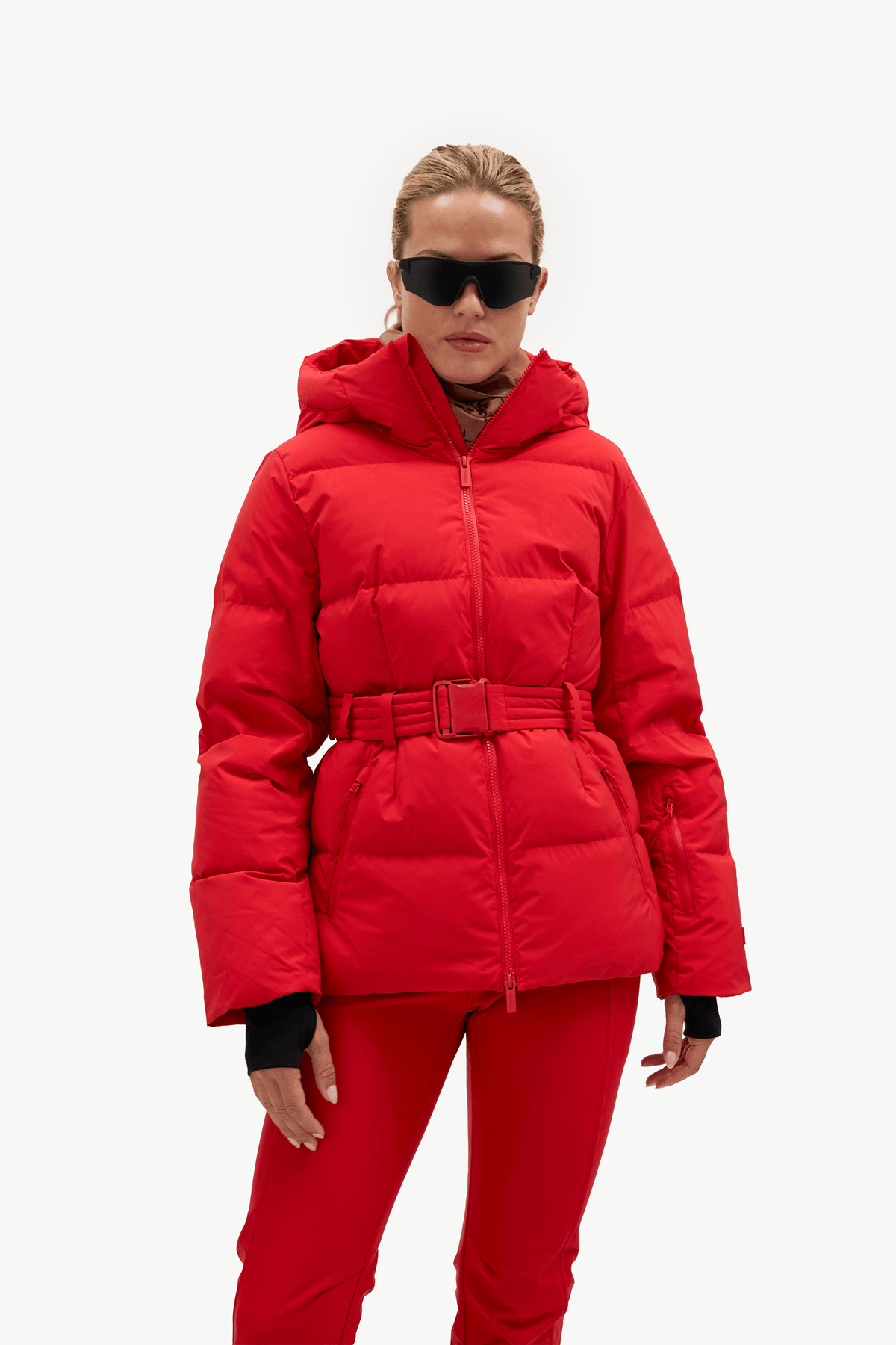 A model wearing a bright red hooded puffer jacket with a matching belt and red pants, styled with black sunglasses, against a plain white studio background.