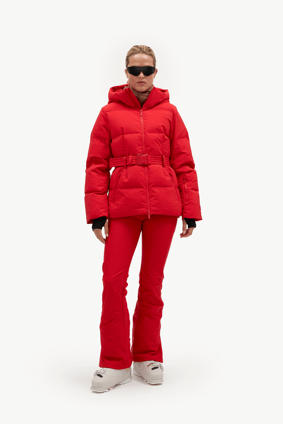 A model wearing a bright red ski jacket with a matching red ski pants and light gray snow boots, standing against a plain white studio background.