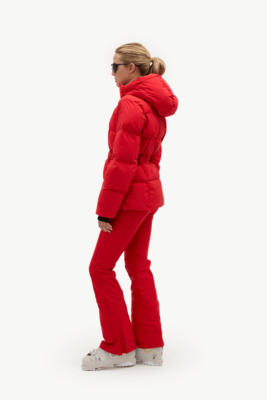 A woman in a bright red puffer jacket and matching red ski pants, shown in profile against a plain white studio background.