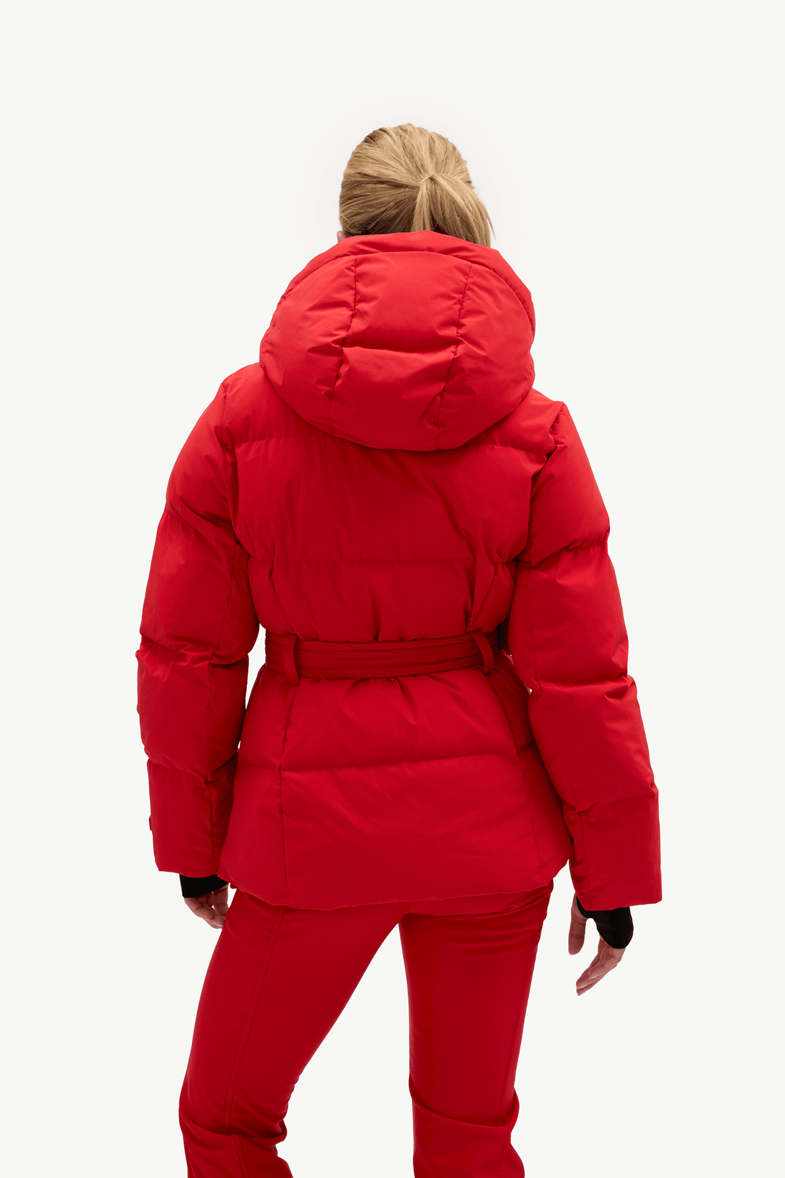 A woman viewed from behind wearing a bright red hooded puffer jacket with a matching belt and red pants against a plain light gray studio background.