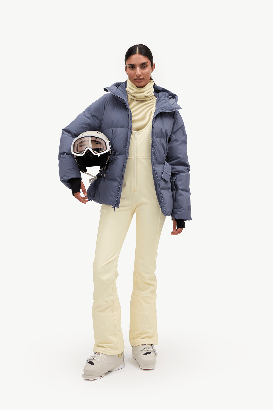 A model stands against a plain white studio background wearing a slate blue puffer jacket over a cream turtleneck and matching cream snow pants, holding a white ski helmet with goggles.