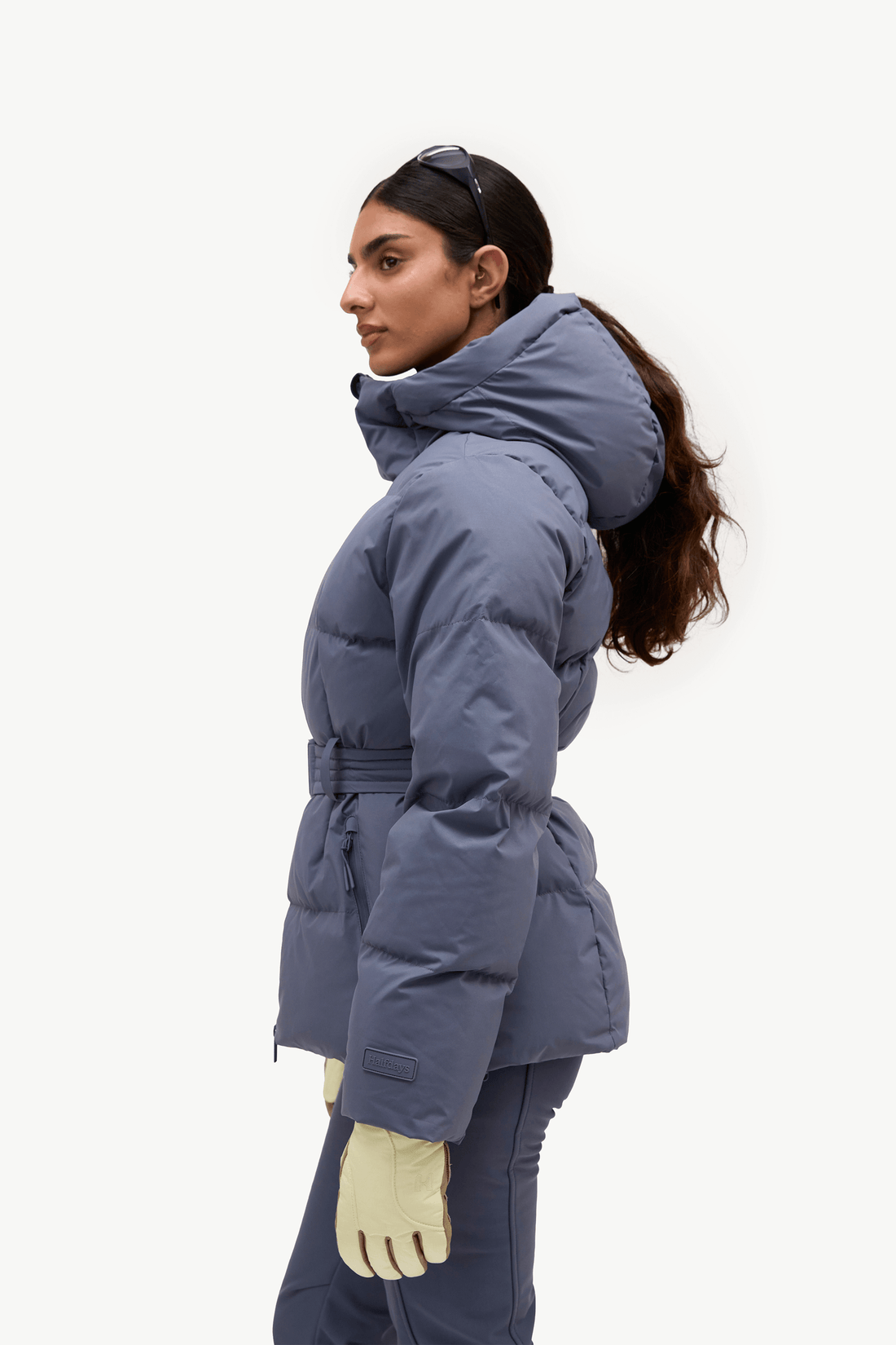 A woman in profile wearing a slate blue puffer jacket with a high collar and waist tie, paired with matching pants and cream gloves, against a plain white studio background.