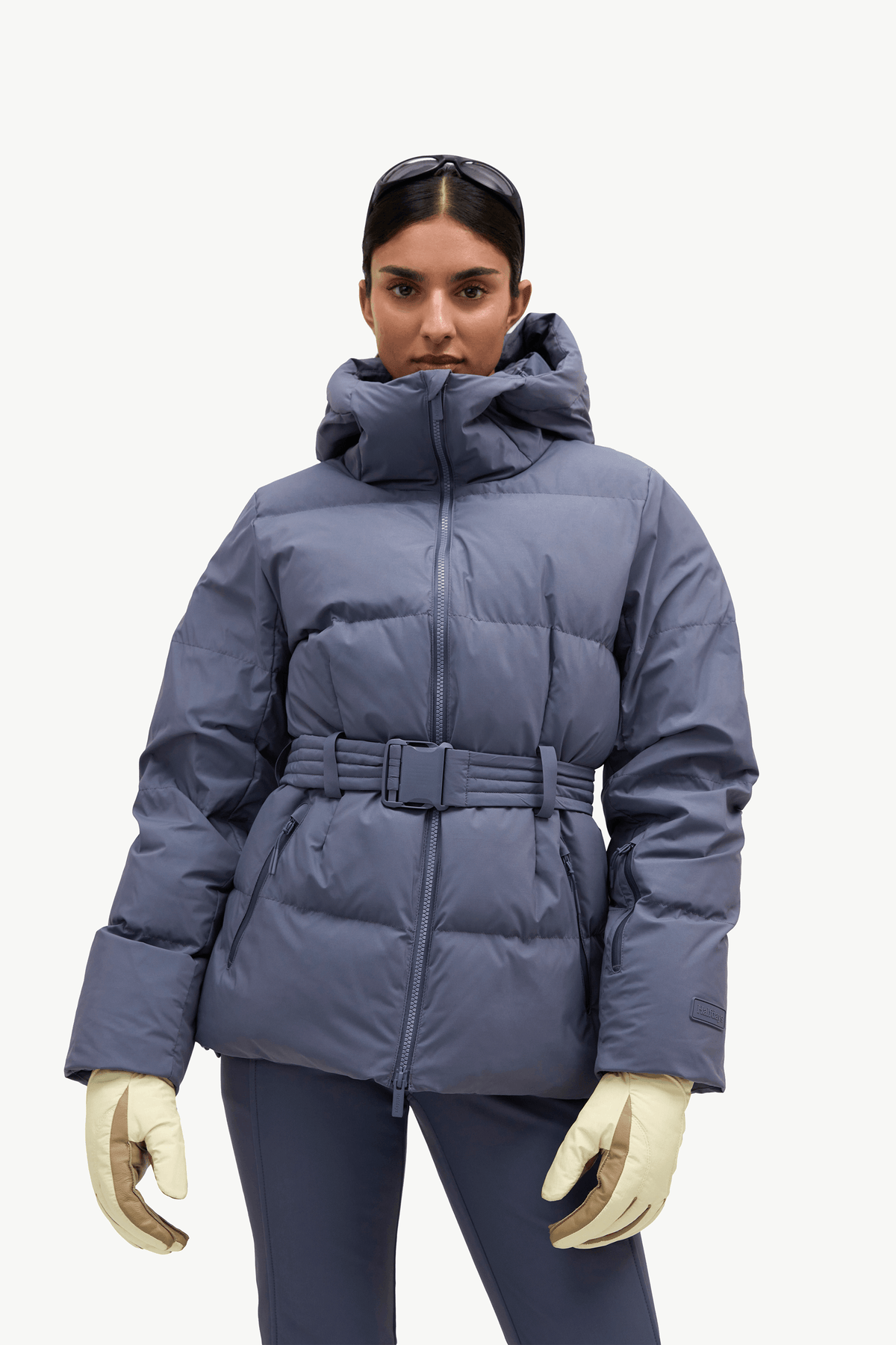 A model wearing a slate blue puffer jacket with a matching belt and hood, paired with dark pants and cream-colored gloves, against a plain light gray background.