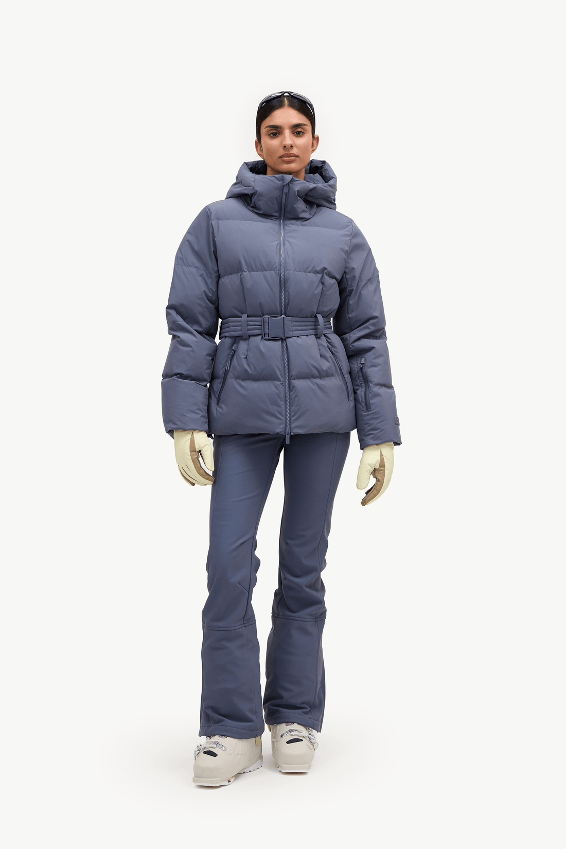 A model stands against a plain white studio background wearing a slate blue puffer jacket with a matching belt, ski pants, gloves, and snow boots.
