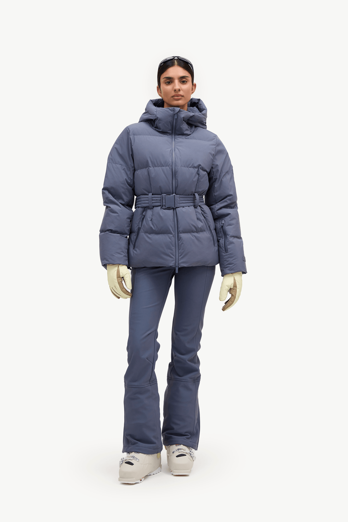 A model stands against a plain white studio background wearing a slate blue puffer jacket with a matching belt, ski pants, gloves, and snow boots.