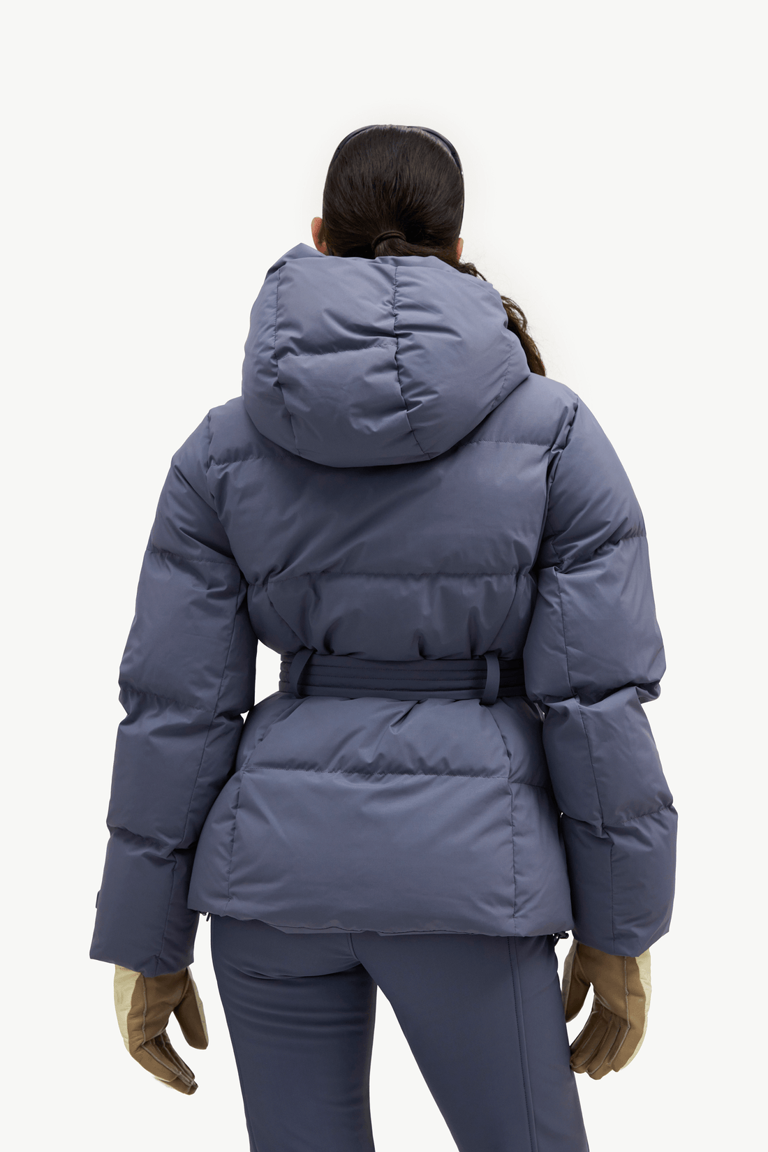 A model shown from the back wearing a slate blue puffer jacket with a belted waist and hood, paired with matching blue leggings and tan gloves, against a plain white background.