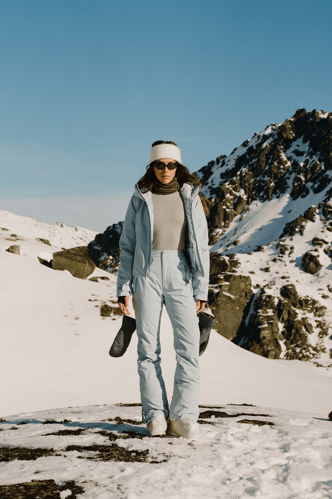A woman stands in a snowy mountain landscape wearing a light blue ski jacket and matching ski pants, a beige top, a white beanie, sunglasses, and holding black ski boots.