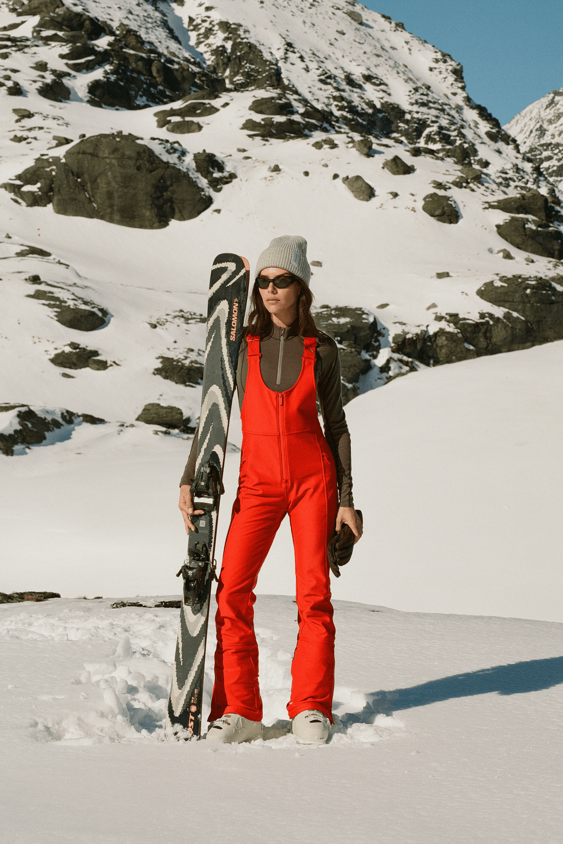 A woman standing in a snowy mountain landscape holds a black and white snowboard. She is wearing a bright red snowsuit, a light gray beanie, sunglasses, and gloves.