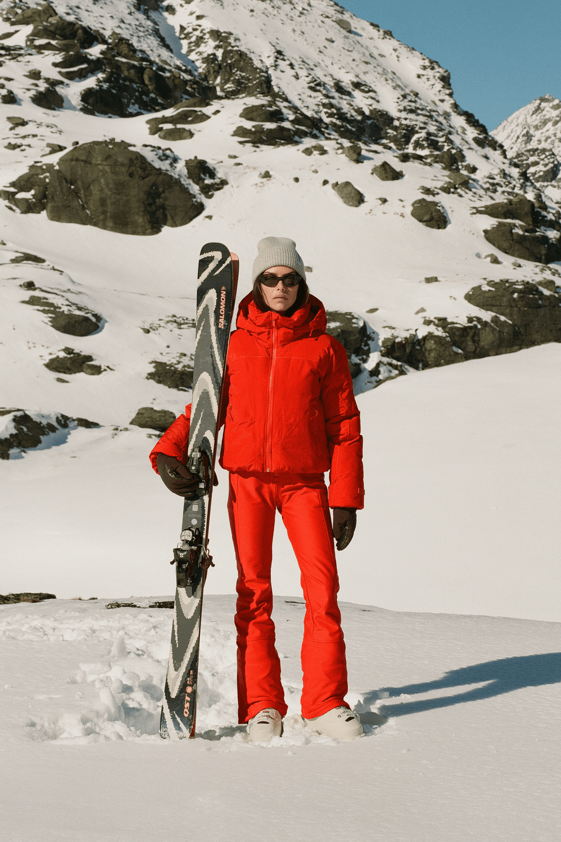 A person standing in the snow wearing a bright red ski jacket and matching ski pants, holding a black and white ski upright, with snowy mountains in the background.