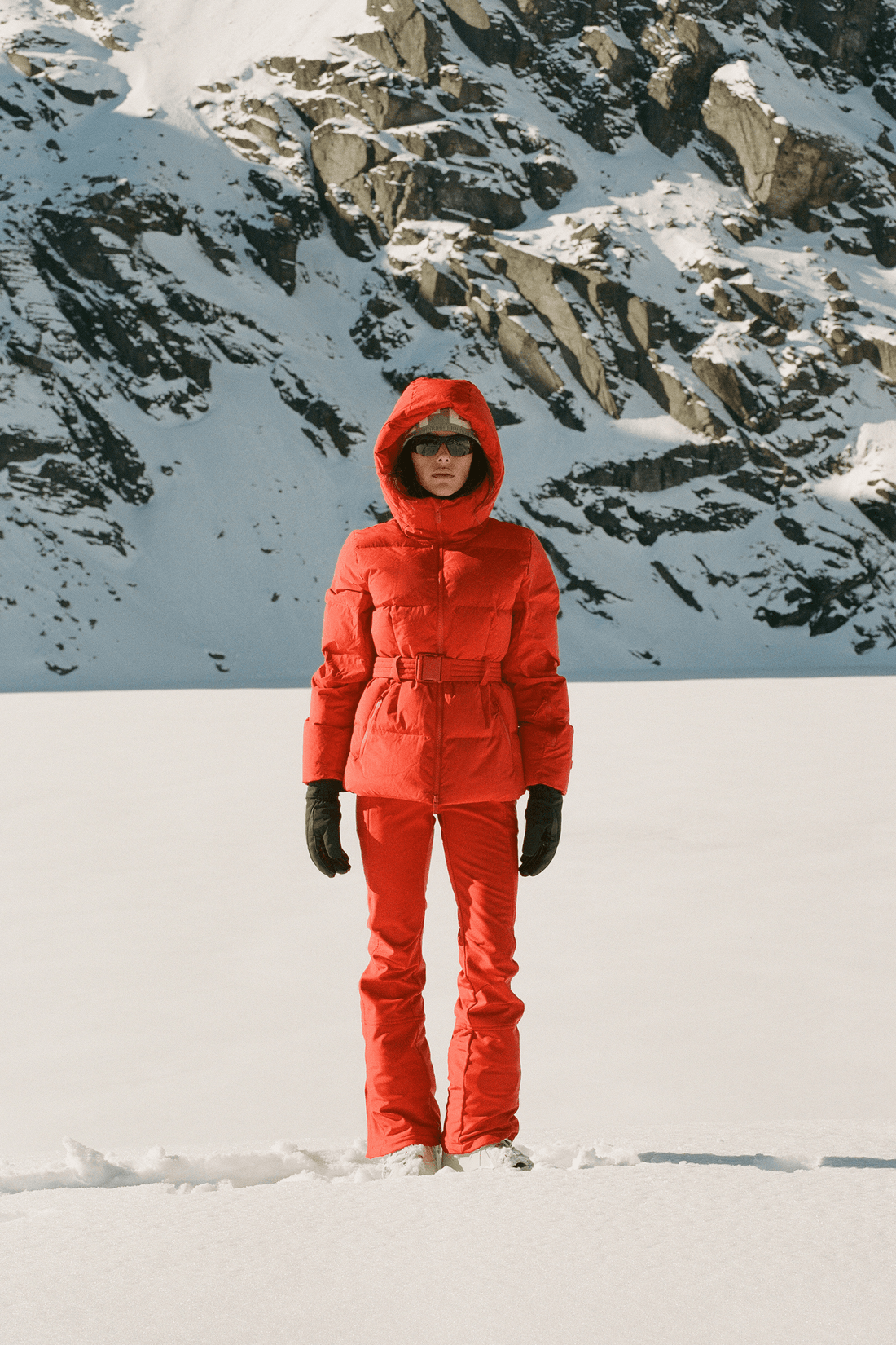 A person standing in the snow wearing a bright red hooded puffer jacket and matching red snow pants, with black gloves, against a snowy mountain backdrop.