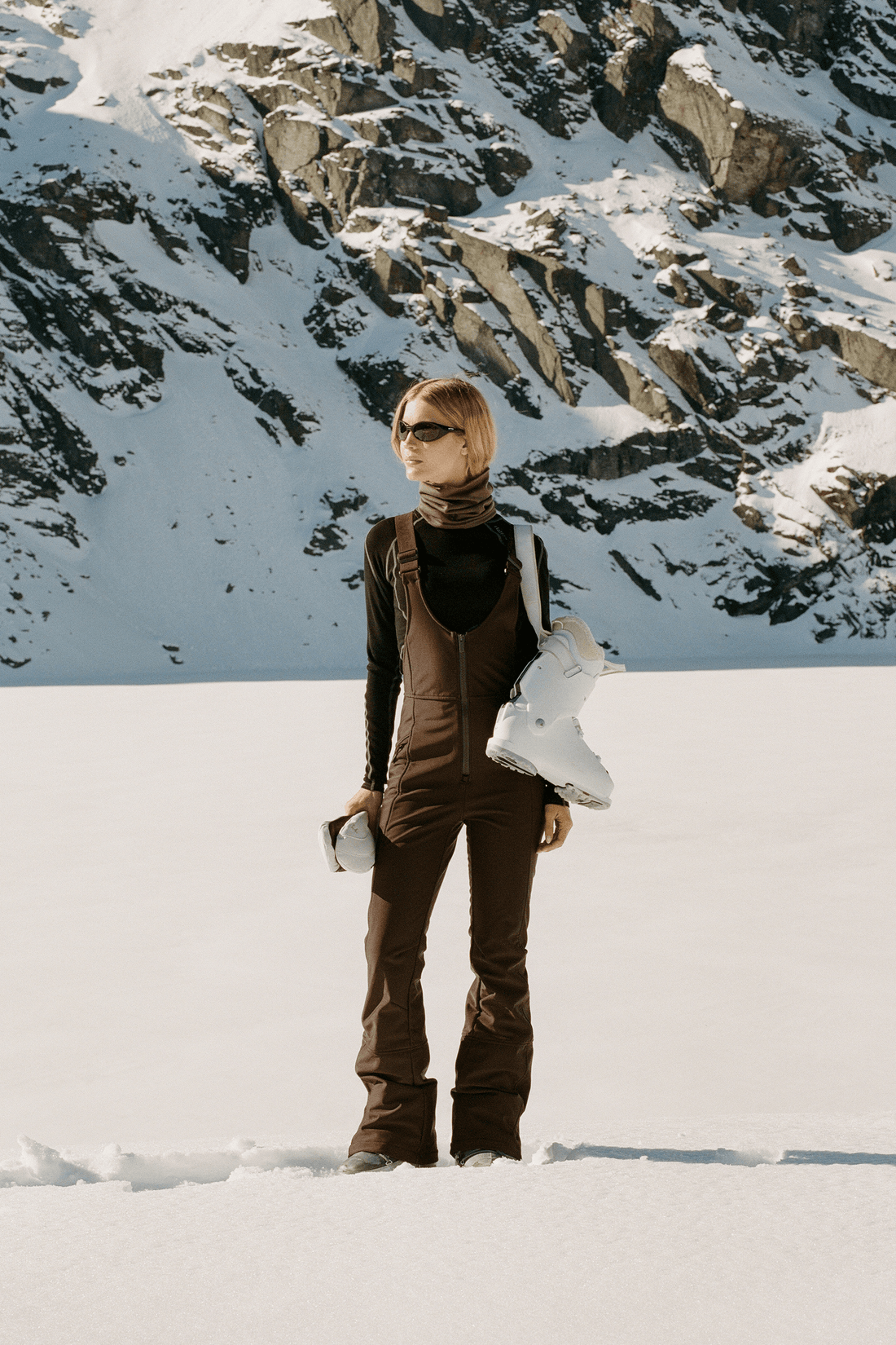 A woman stands in a snowy mountain landscape wearing brown snow pants and a black turtleneck, with a white snowboard on her shoulder.
