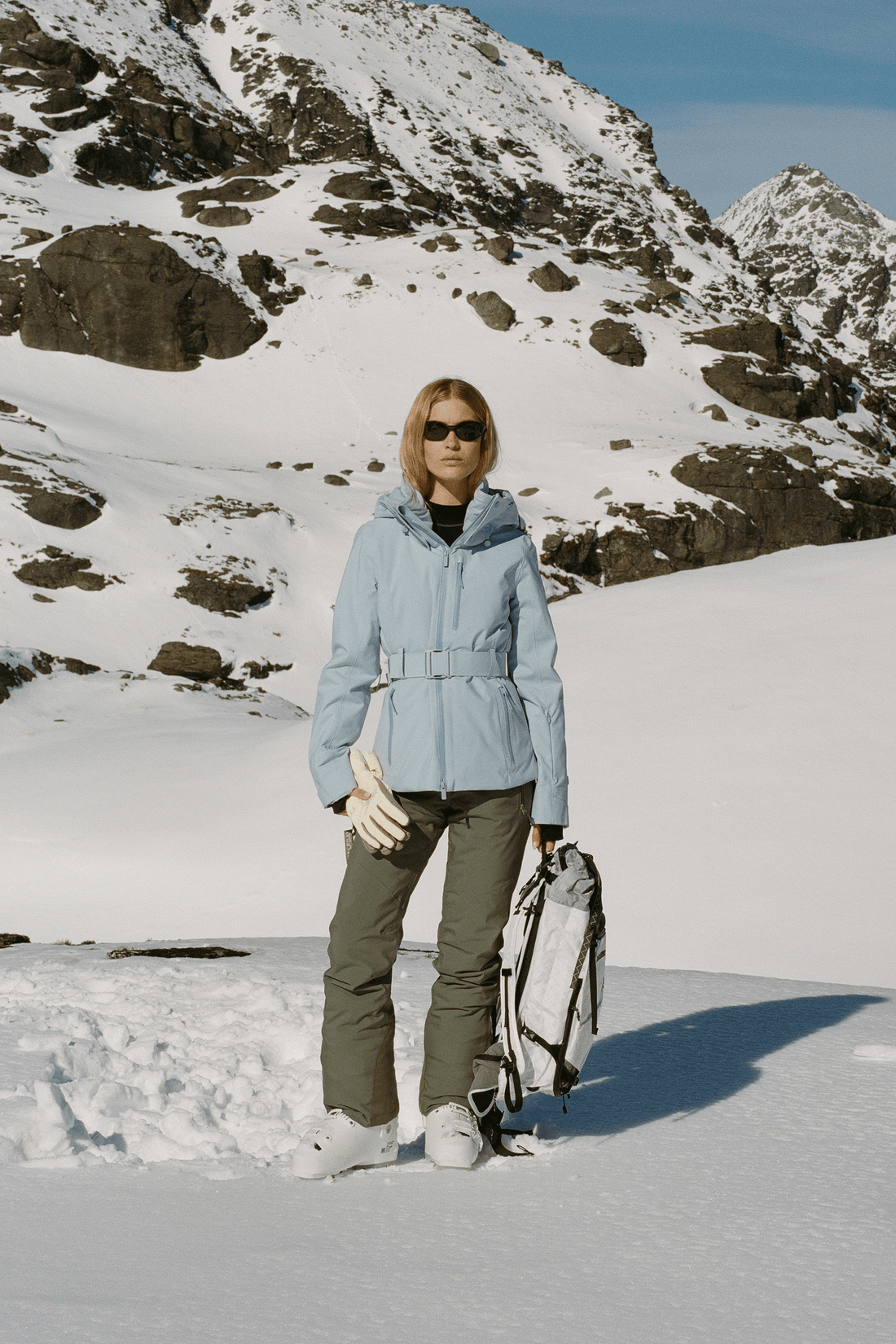 A woman stands in a snowy mountain landscape wearing a light blue hooded jacket, olive green pants, gloves, and a backpack.