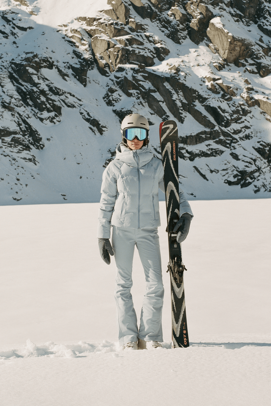 A person standing in the snow wearing a light gray ski outfit, helmet, goggles, and gloves, holding a black and white ski in front of rocky, snow-covered mountains.