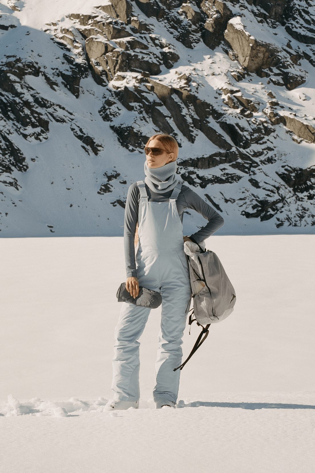 A person standing in snow wearing a light blue snowsuit, gray long-sleeve top, gray neck gaiter, sunglasses, and carrying a gray backpack, with rocky mountains in the background.